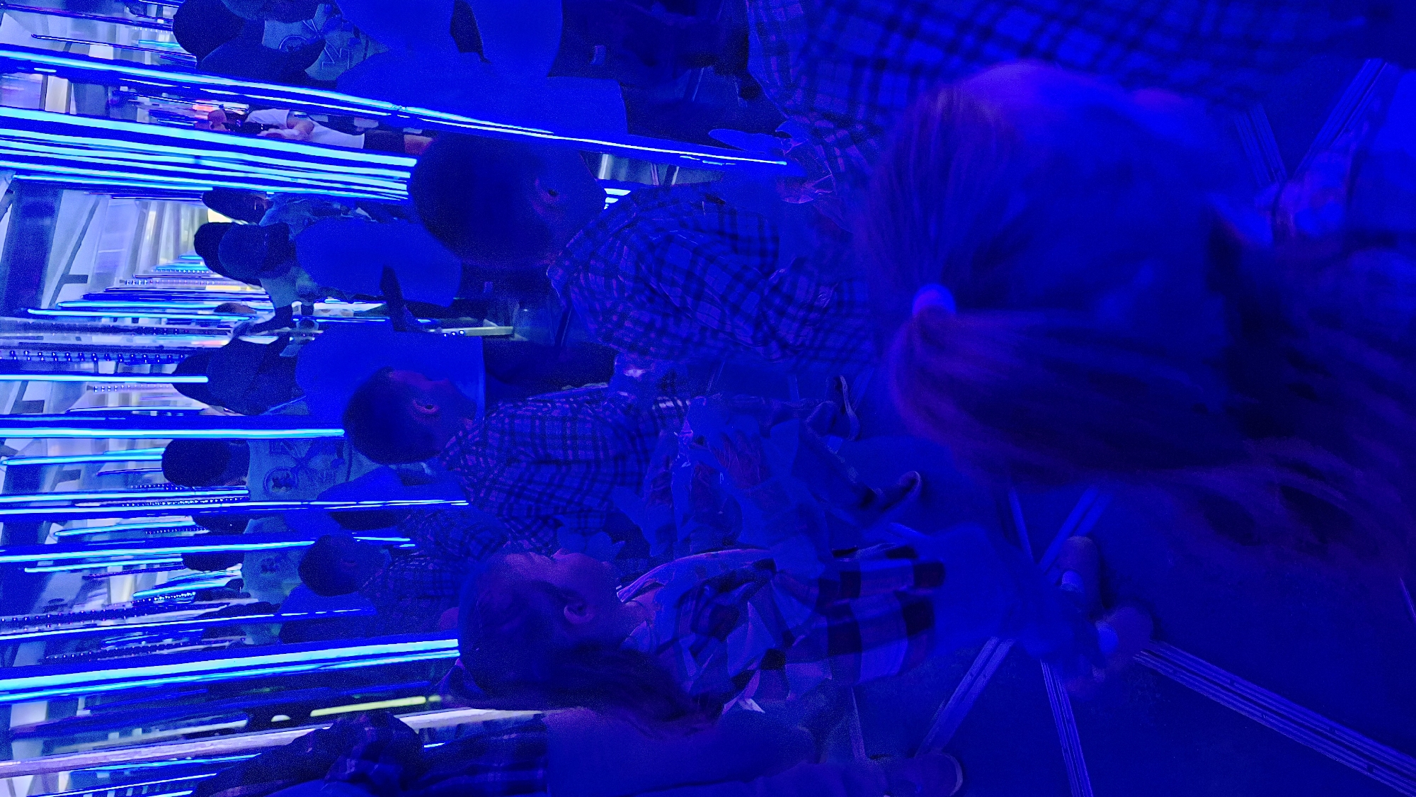 The photograph depicts a mirrored installation art piece filled with the reflections of people, potentially taken indoors. The entire image is lit with bright blue light, predominantly from horizontal light bars running across the scene. Multiple faces and figures are visible due to the reflections from the mirrors. At the bottom of the frame, a person lies down in a plaid shirt. The overall aesthetic of the image is intensely blue with a complex interplay of reflections.