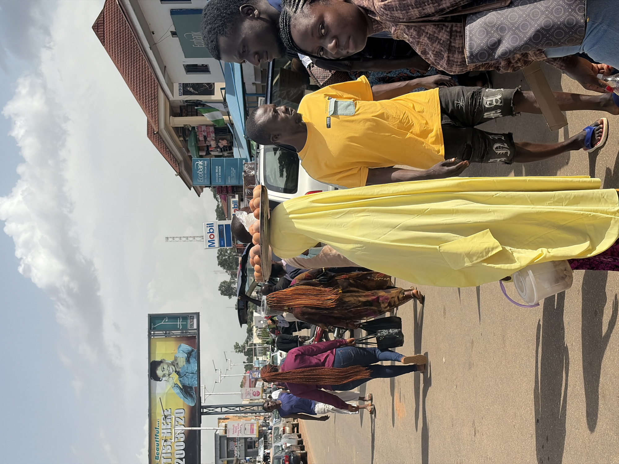 The image depicts a street scene in Jos, Nigeria. In the foreground, a person wearing a bright yellow garment is partially visible, carrying a tray of eggs on their head. To the right, a man in a yellow t-shirt and ripped denim shorts stands looking to the left. Next to him, a woman with dark braided hair in a brown patterned top is also looking towards the left. Behind them, other individuals are walking along the street. On the left side of the image, a large billboard advertising "Beautiful... ARTIST HERE" and "2005 0720" is visible, along with the Ecobank logo and a sign for Mobil. The street is paved, and several cars are parked or driving in the background. The sky is partly cloudy.