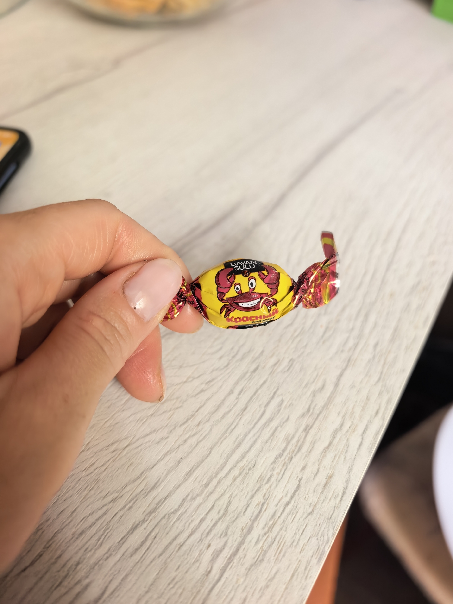A hand holds a wrapped candy. The wrapper is yellow with a red crab cartoon on it. The words "BAYAN SULU" are written in black above the crab. Below the crab, the word "КРАСНЫЙ" is written in red, followed by the word "карамель" in smaller black letters. The wrapper is twisted at both ends. The background includes a light-colored wooden surface and other objects that are out of focus. There is no explicit visual information to confirm the location in Sterlitamak, Russia.