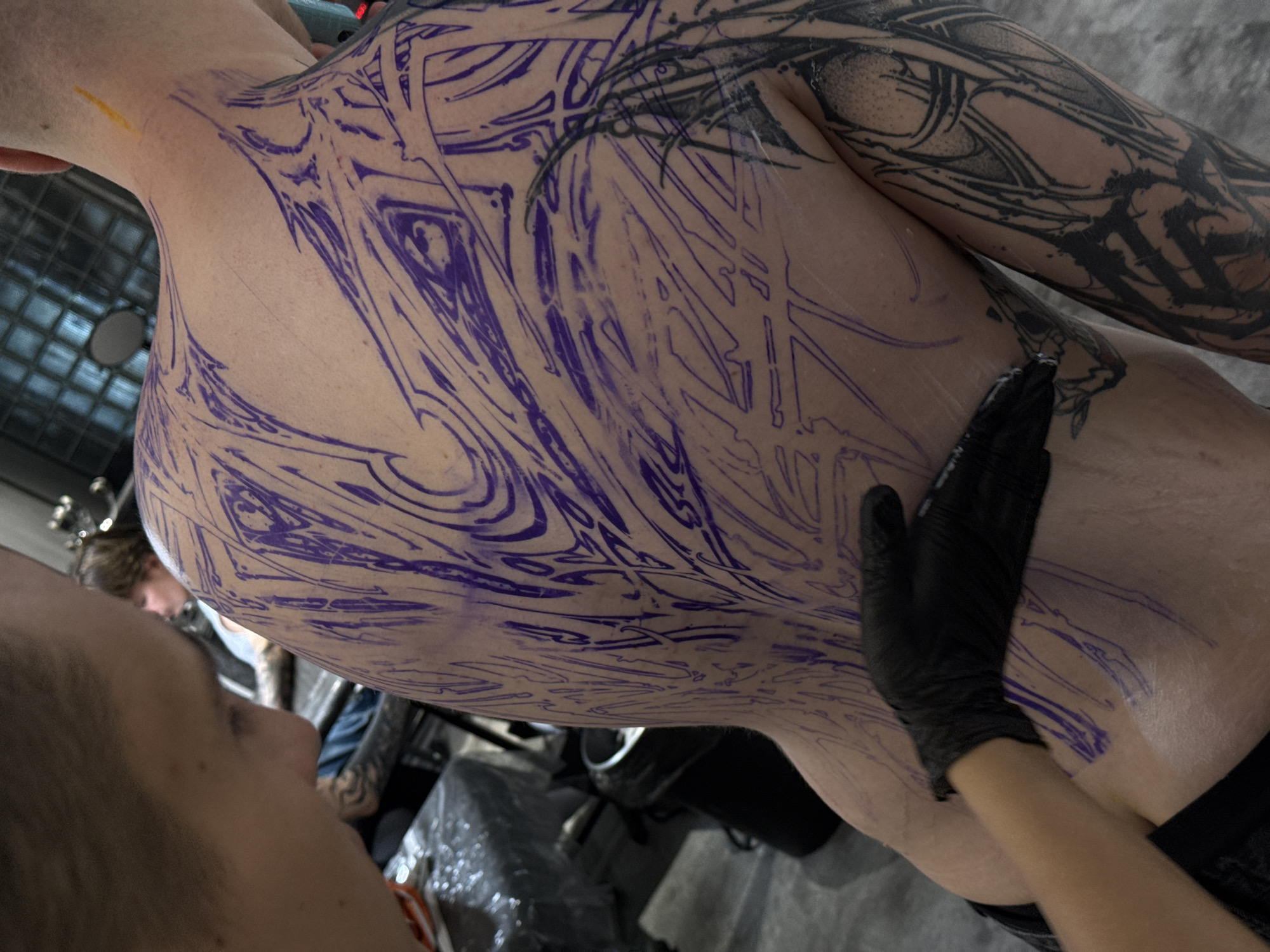 A person's back and arm are shown, covered with a large tattoo design in purple and black ink. The purple lines appear to be a stencil for the tattoo, while the arm already has a completed black ink tattoo. A gloved hand is pressing on the lower back, possibly applying or smoothing something. The scene likely depicts the tattoo application process. Based on the style and subject matter, it is potentially in Moscow, Russia, but that cannot be confirmed with certainty from the image.