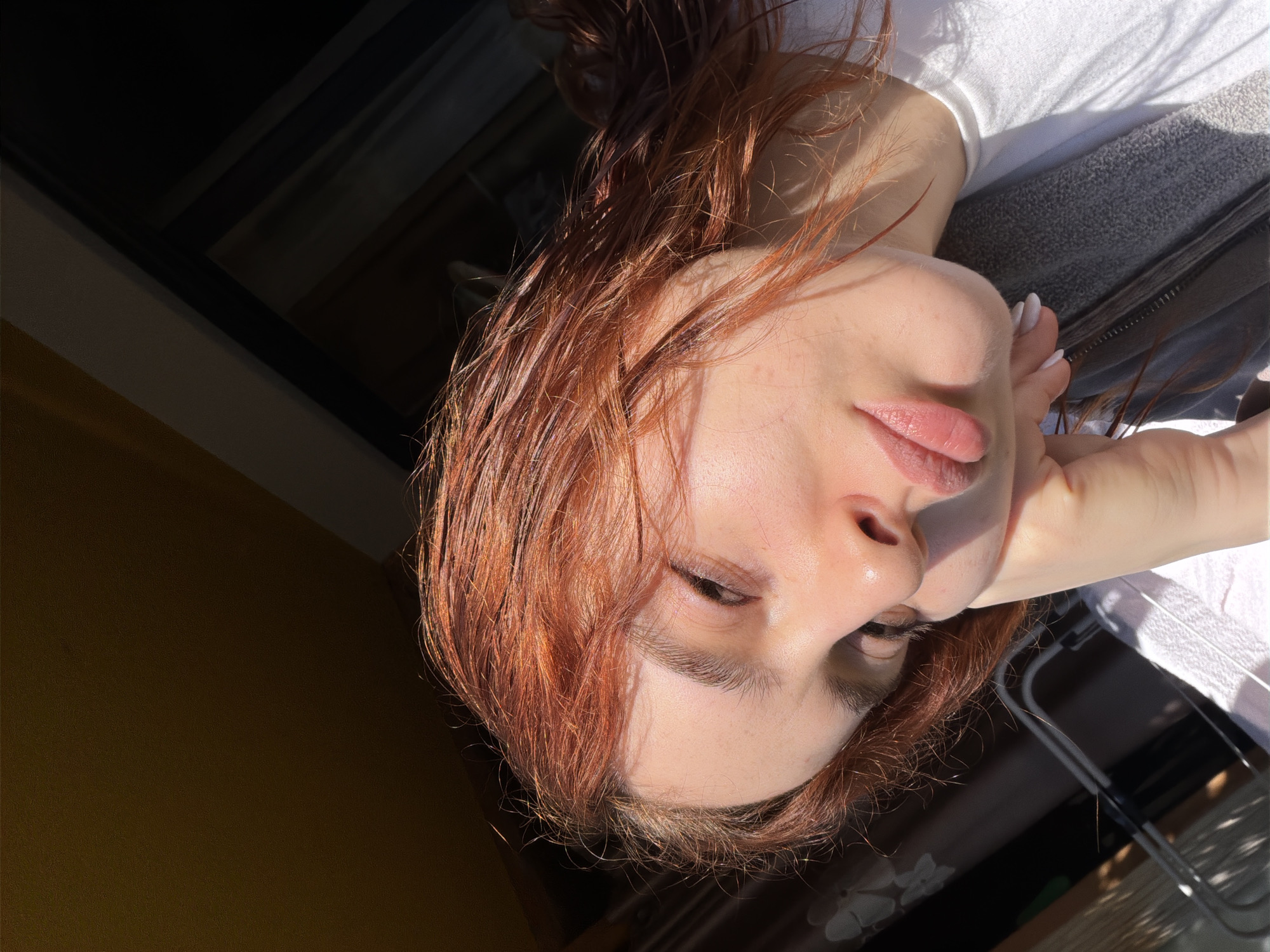 A close-up, rotated image shows a person with red hair, fair skin, and closed eyes. The person is wearing a white shirt and leaning against a grey object, possibly a jacket. The location appears to be indoors, near a window. Based on the lighting and shadows, the environment may have strong sunlight streaming in. Given the limited information, specifically mentioning the location in the city of Strasbourg, France is not feasible as it requires additional clues within the image.