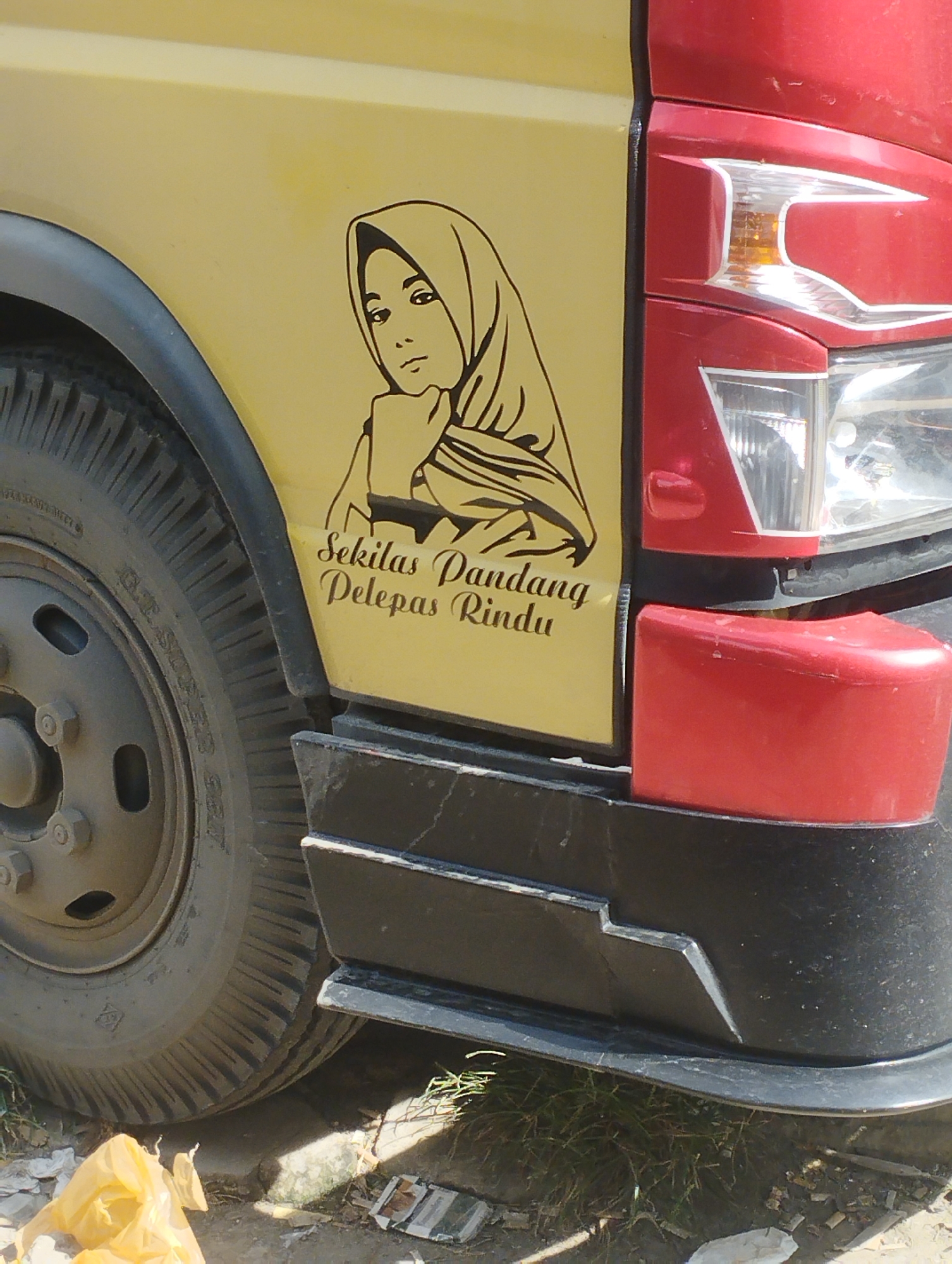 The image shows the front of a vehicle, specifically the wheel, part of the door, and headlight area. The door is yellow and features a black line drawing of a woman wearing a headscarf, along with the Indonesian phrase "Sekilas Pandang Pelepas Rindu." The vehicle's front is a dark red color. There is a tire partially visible on the left side of the frame. The vehicle appears to be parked on a street or in an outdoor area. The location in the city of Lhokseumawe, Indonesia is not visually verifiable.