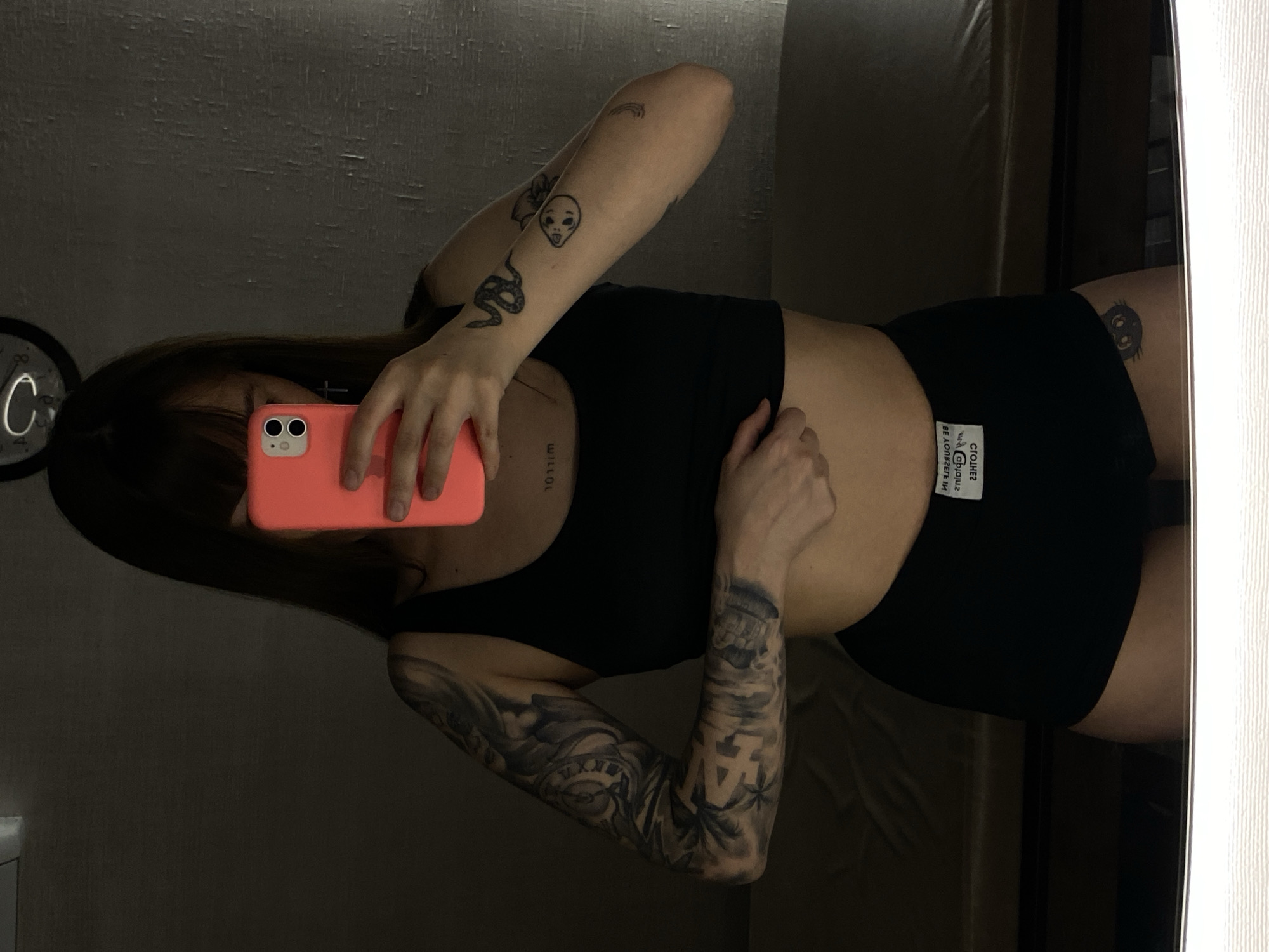 A mirror selfie captures a person indoors, in what appears to be a dimly lit room in Votkinsk, Russia. The individual, seen from the waist up, is female, with long dark hair partially obscuring her face as she holds a bright coral/orange iPhone to take the picture. She is wearing a black athletic crop top and matching black shorts.
Her body is extensively tattooed. Her left arm features a full sleeve with intricate black and grey designs, including a large "LA" lettering, clock faces with Roman numerals, and what appear to be clouds or foliage. On her right arm, closer to the bicep, are smaller distinct tattoos: a coiled snake, an alien head, and a small rainbow. Faint, inverted text that reads "WILLOW" is visible on her left ribcage. On her right upper thigh, a dark, cartoon-style owl-like or monster-like face is tattooed.
The background includes a dark, possibly textured wall. A circular clock face, partially visible on the left, shows a large letter "C" or "G." On the white tag of her black shorts, inverted text reads "HE REMEMBERS MY SMILES," along with smaller, possibly brand-related text like "CROL." The right side of the frame shows a light-colored wall or mirror border. The overall lighting suggests evening or night, or a space with intentionally low illumination.