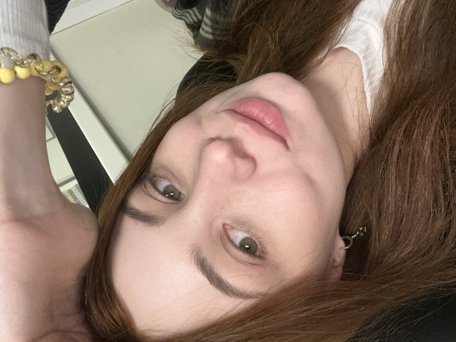 A woman lies with her head tilted, showcasing hazel eyes and pink lips. Her auburn hair frames her face, and she wears a silver dangling earring. A gold bracelet with yellow beads adorns her wrist. The background includes a white surface and hints of black clothing. The composition is close-up and focused on the woman's face.