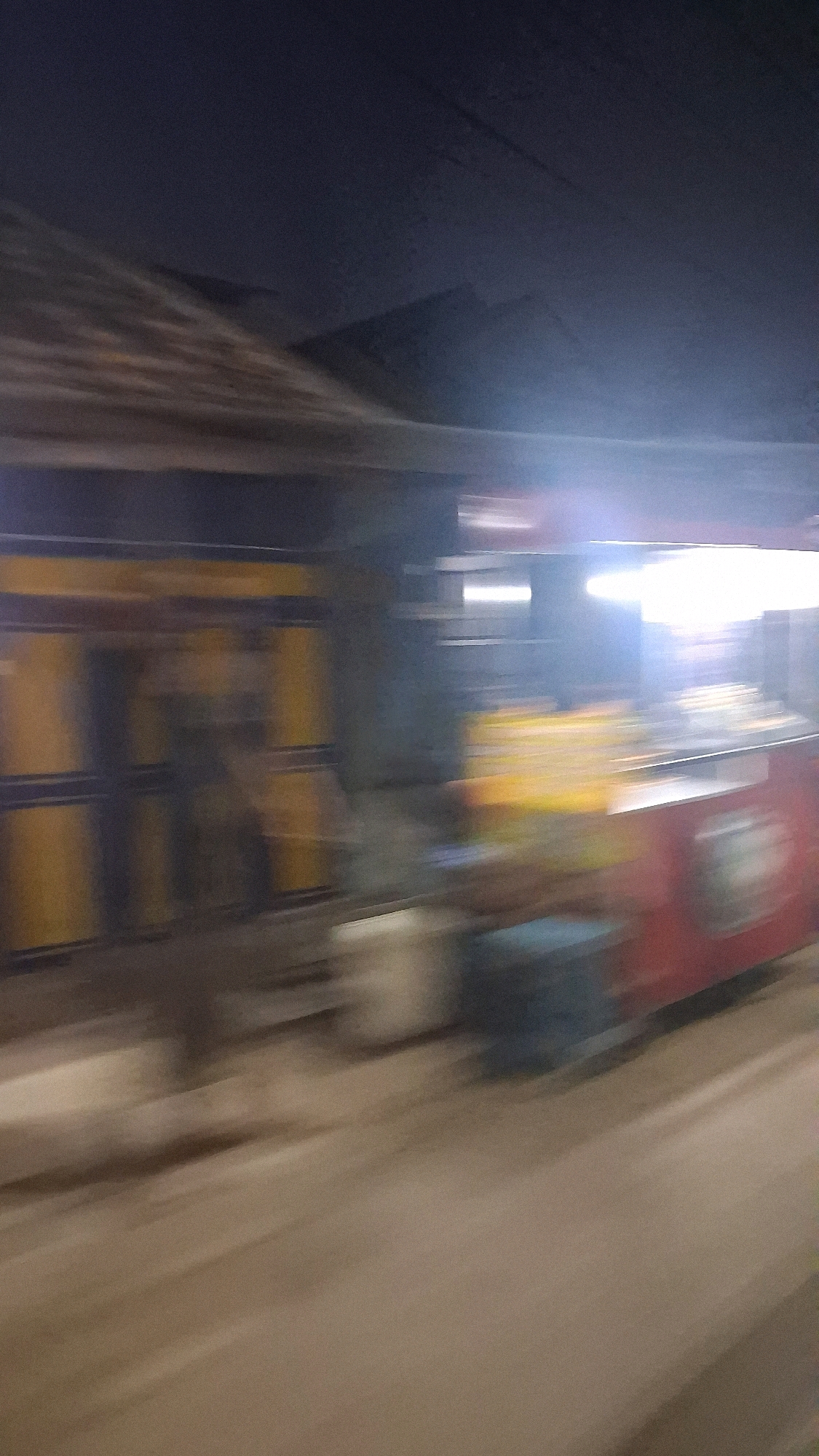 The image captures a blurred street scene at night, likely in Jogotirto, Indonesia, due to motion blur. On the left, a building with a weathered, dark roof is visible, featuring yellow and dark blue panels, possibly a shopfront or dwelling. To the right, a brightly lit food cart or stall is in motion, with a red base and a counter displaying illuminated yellow and white items, suggesting food being prepared or displayed. A dark blue container is situated near the food cart. The background consists of dark, indistinct shapes of hills or mountains against a night sky. The ground appears to be a paved surface, likely a road, with a lighter shade indicating the presence of streetlights or other illumination. The overall impression is one of a dynamic, transient moment, possibly from a moving vehicle, capturing a slice of nocturnal street life.