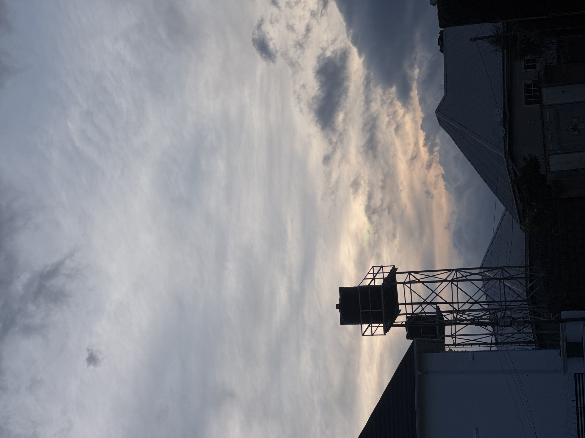 An outdoor, eye-level, medium shot captures the scene of a water tower and rooftops silhouetted against a dramatic cloudy sky at dusk. The sky is filled with a mix of dark grey and lighter grey clouds, with patches of muted yellow and pink hues suggesting the last light of the sun breaking through. In the foreground, the sharp, dark angles of two house rooftops are visible. To the left, the white side of a building is partially visible. The central focus is a tall, dark water tower with a rectangular tank at the top, supported by a metal lattice structure. Another, smaller water tank is visible below it, also on a metal frame. The overall atmosphere is one of a quiet, urban twilight.