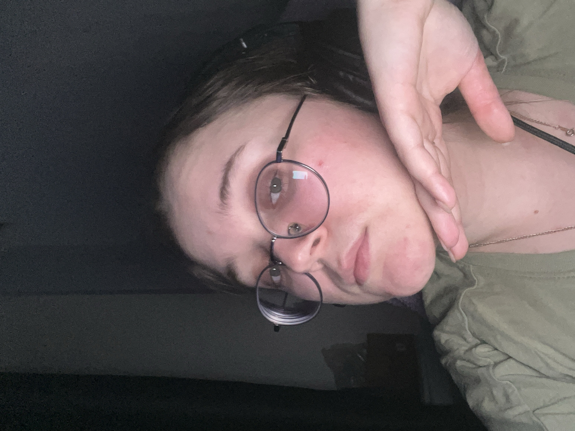 A close-up portrait-style image, taken in Pushchino, Russia, features a young person indoors. The individual, seen from the chest up, has their head tilted and looks directly at the camera with a neutral to slightly pensive expression. They wear round, thin-rimmed glasses and a plain olive-green t-shirt, with a thin golden chain necklace visible around their neck.
The person's right hand is raised, with the palm facing the viewer, partially covering their left cheek and jawline, and resting near their ear. The index finger of this hand shows some redness or irritation. A small red blemish is noticeable on the person's forehead, and there is also some redness on their cheek.
The lighting is dim, suggesting an evening or low-light indoor setting, and casts soft illumination on the person's face. The background is dark and indistinct, reinforcing the indoor environment. Reflections, possibly of a light source or the camera itself, are visible in the lenses of the glasses. No visible text is present in the image.