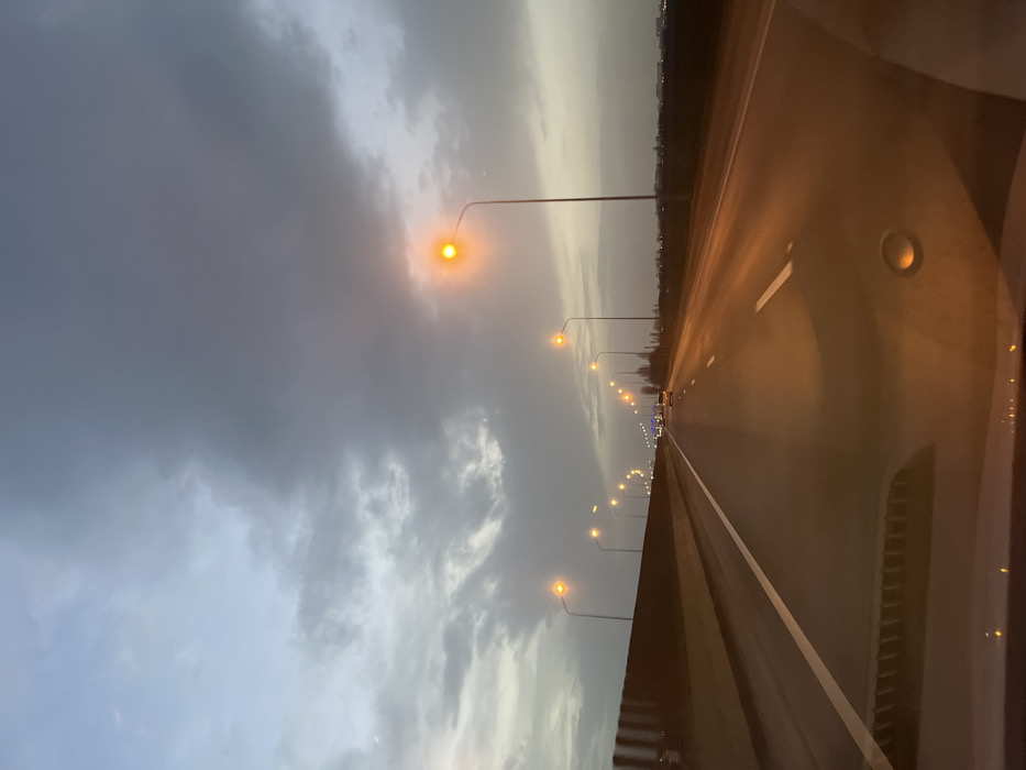A photograph depicts an empty highway at dusk. Streetlights line the left side of the road, their orange glow contrasting with the grey, cloudy sky. The highway surface reflects the light. A white line marks the edge of the lane. The image appears to be taken from inside a vehicle, with some interior elements visible on the right side of the frame. The overall mood is serene and atmospheric.