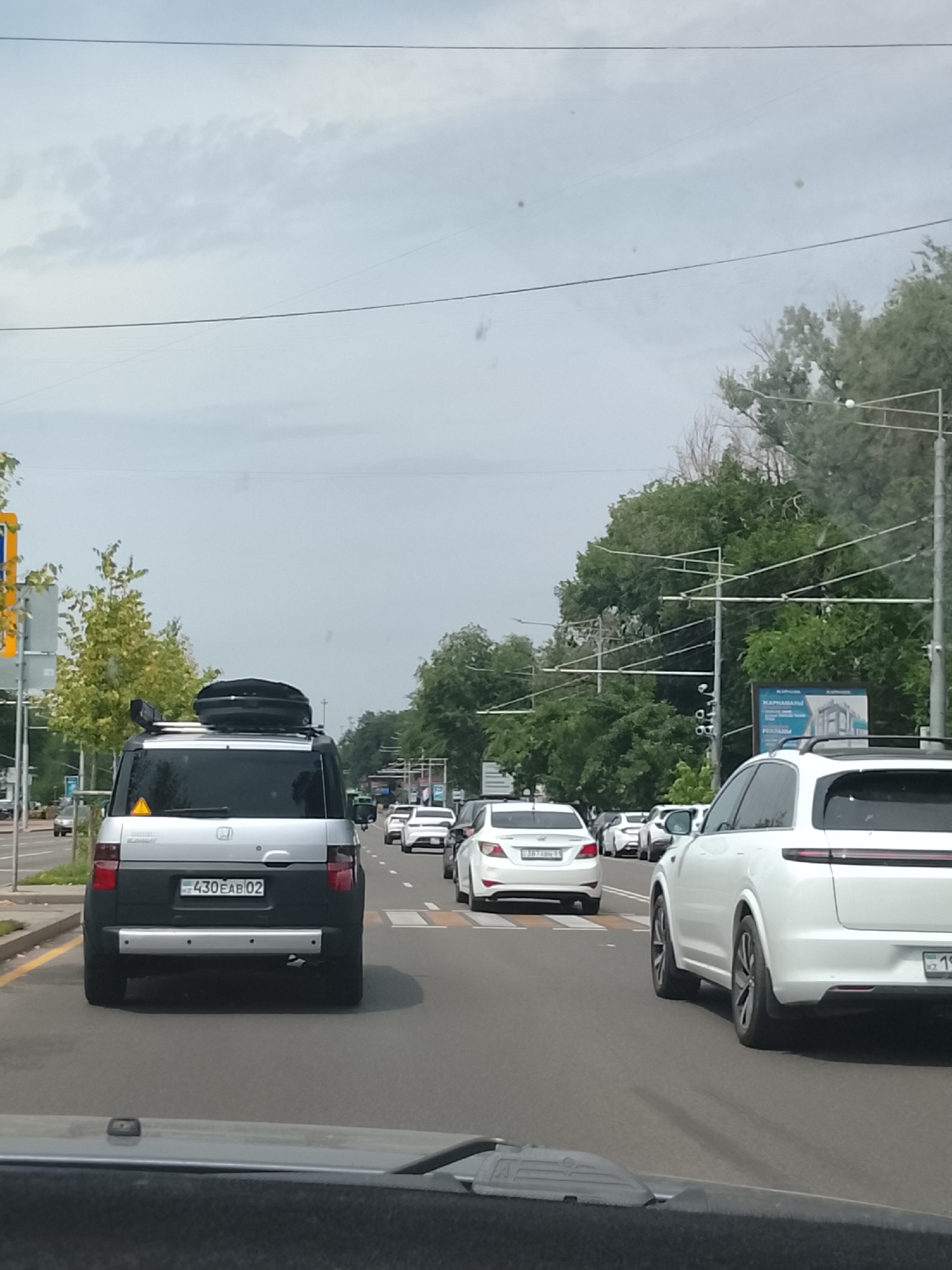 The image shows a view from a car in traffic on a city street in Almaty, Kazakhstan. The foreground shows the dashboard of the car from which the photo is taken. In front, a silver Honda Element with a black roof box is in the lane. The license plate of the Honda Element is visible and reads "KZ 430 EAB 02". Further ahead, several white cars are visible, including a white Hyundai Accent with license plate "387 ABN 11". The road has multiple lanes, and traffic appears to be moderate. Trees line the right side of the road, and utility poles with wires are visible overhead. A billboard with images and text in Russian is also present on the right. The sky is overcast.