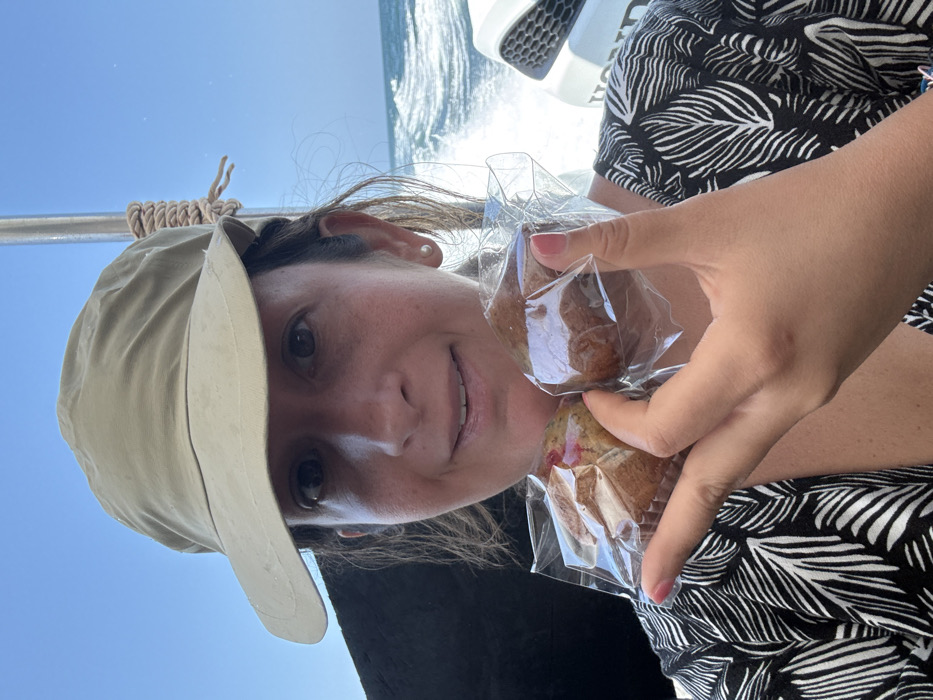 An image taken in Pak Nam, Thailand, depicts a female individual on a moving boat under a clear blue sky. The woman, who appears to be of middle age, wears a light tan bucket hat and a dark garment with a light leaf pattern. She exhibits a slight smile, revealing her teeth, and has a single white pearl earring visible in her right ear. Her left hand, with light pink painted fingernails, holds two individually plastic-wrapped muffins prominently in the foreground. One muffin, positioned to the left, appears to contain red fruit pieces, while the other is a plain brown variety. In the background, turbulent water and white wake are visible on the right, indicative of a vessel in motion. A white outboard motor bearing the "HONDA" brand logo is partially visible on the far right. A vertical metal pole wrapped with light-colored rope is positioned to the right of the woman's head, suggesting boat infrastructure.