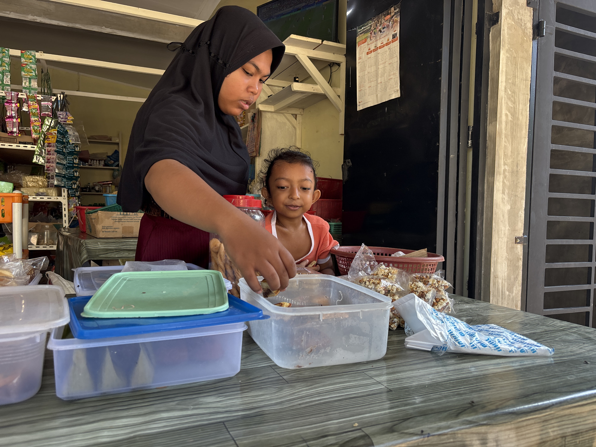 In Lhokseumawe, Indonesia, a woman wearing a black hijab and a maroon skirt stands behind a table. She is leaning forward, her right hand reaching into a clear plastic container filled with food. Beside her, a young child in an orange shirt watches. On the table are several plastic containers, one with a green lid and another with a blue lid stacked on top, as well as bags of snacks. The background shows shelves stocked with goods, a calendar, and a metal gate. The scene appears to be an outdoor market or small shop.