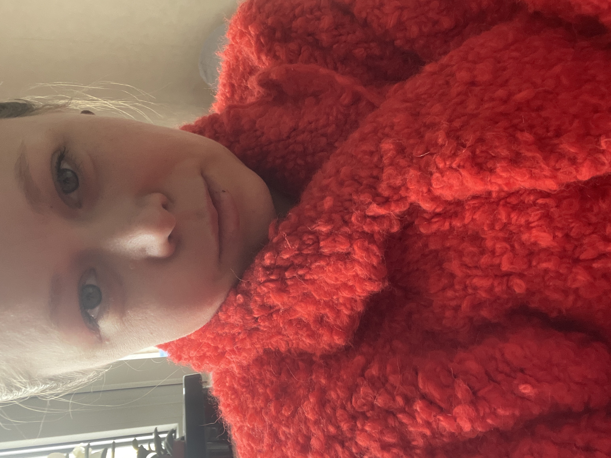 A selfie of a fair-skinned person with light blonde hair. They are wearing a bright red, textured, possibly wool or fleece garment. The background appears to show a window with closed blinds, and potentially a shelf below. The person's face is angled towards the camera, with a slightly pensive expression. There is no readily apparent evidence linking this image to the city of Chaikovsky, Russia.