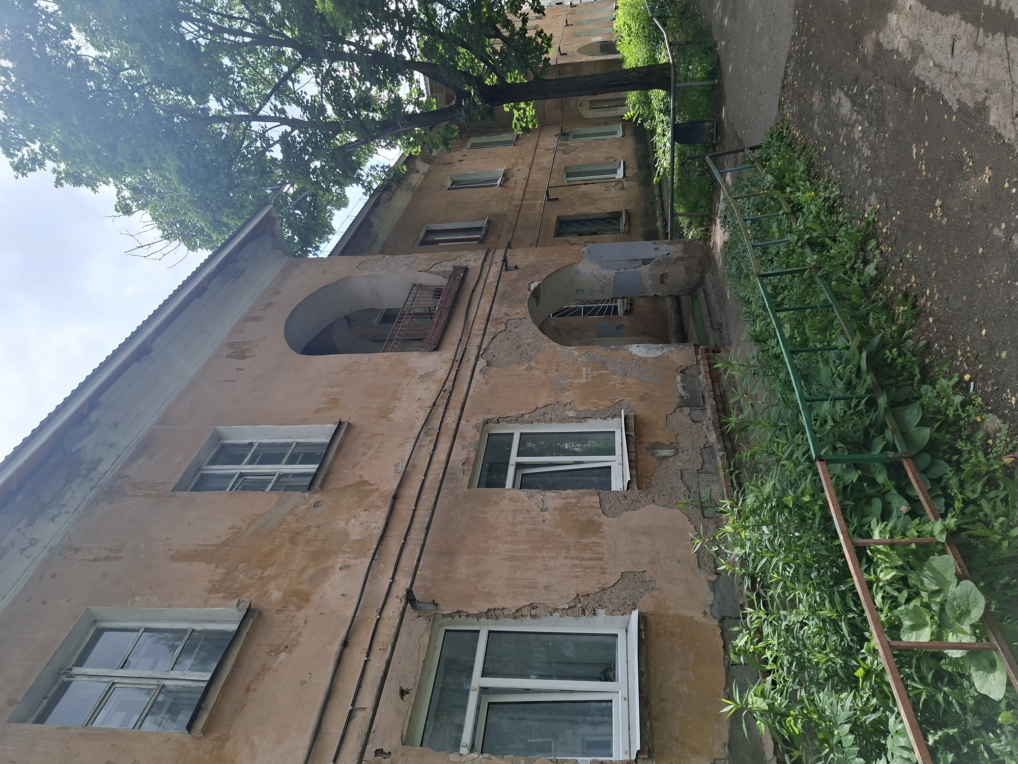 The image depicts the exterior of an aging apartment building, likely in Izhevsk, Russia, given the context provided. The building's facade is a faded, peeling peach color, showing signs of significant wear and tear, with large patches of plaster missing, exposing the brickwork beneath. There are several windows visible, some with white frames and glass panes reflecting the sky and trees. One window on the left appears to be a modern replacement with clean white frames, contrasting with the older, weathered appearance of the building.
The building has several architectural features, including arched doorways and a balcony with a wrought-iron railing. Electrical conduits and wires run along the exterior wall. A large, mature tree with lush green foliage stands to the right of the building, its branches reaching towards the sky.
In the foreground, a rusty metal fence with green vertical bars separates the building from a patch of overgrown green vegetation, consisting of various weeds and plants. Beyond the fence, a paved walkway or road is visible, with scattered leaves and debris. The sky is overcast with a bright, diffused light, suggesting it is daytime. There are no people or animals visible in the image. The scene conveys a sense of neglect and age, characteristic of older urban residential areas.