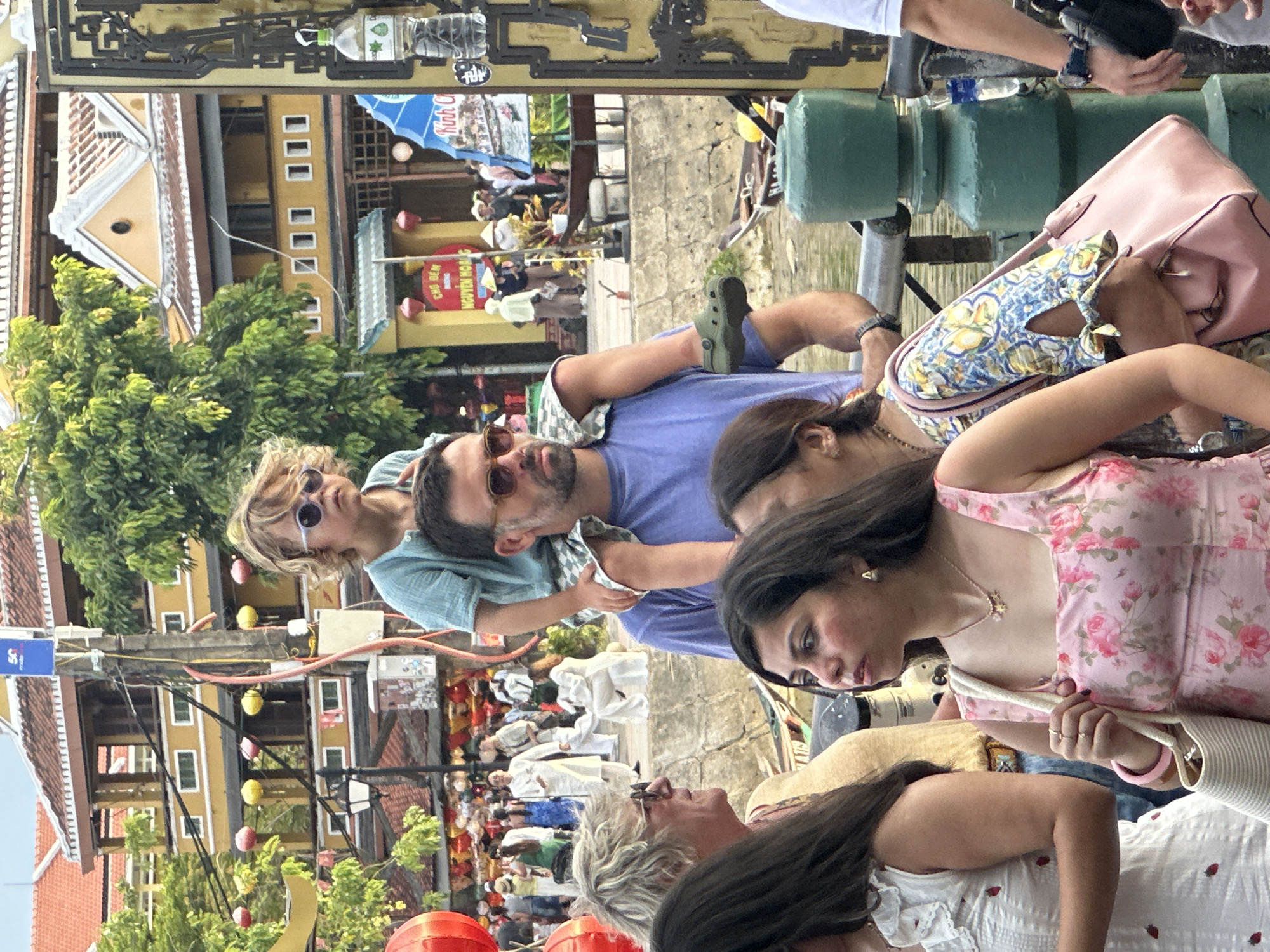 This media file captures a moment with several people in what appears to be a bustling outdoor marketplace or street in Hội An Ward, Vietnam. The subjects are a mix of adults and children, some of whom are interacting. One adult, wearing sunglasses and a blue shirt, is looking towards the camera, while a woman beside him, with dark hair, is also looking away from the camera. A child with blonde hair and sunglasses is looking up, and another person in a pink floral dress is looking down. The scene is filled with various objects, including bags, clothing, and decorative elements like strings of lanterns or lights.
The background reveals buildings with distinct architecture, characteristic of Southeast Asian cities, with yellow facades and tiled roofs. There are also glimpses of trees and what looks like a waterway or canal, further suggesting a riverside setting. The presence of many people in the background suggests a lively atmosphere, possibly a festival or peak tourist hours.
Notable details include the time of day, which appears to be daytime, possibly late morning or afternoon, given the lighting. The weather seems pleasant, with bright sunshine. The overall emotion conveyed is one of casual observation and interaction, with a mix of candid and posed expressions among the individuals in the foreground.
Visible text in the image includes a blue sign with what appears to be Vietnamese characters, and some lettering on a yellow building that is partially obscured. The presence of Vietnamese text and the architectural style strongly indicate the location as Hội An Ward, Vietnam, a well-known tourist destination. The activity appears to be a sightseeing or leisurely stroll through the vibrant streets of the town.