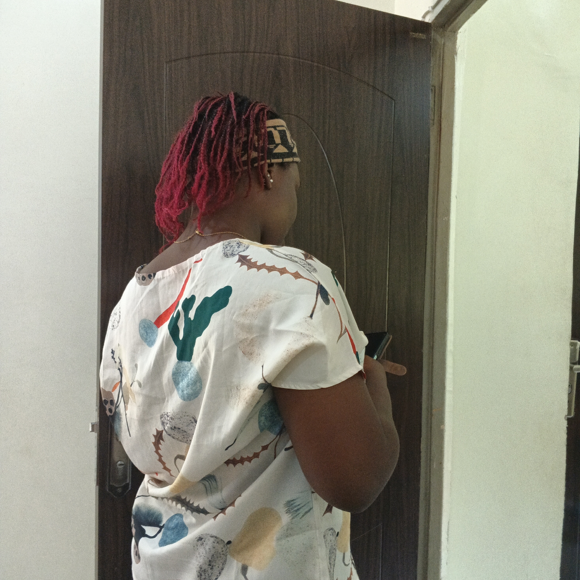 The image depicts a rear view of a Black person standing near a doorway. The person has bright red dreadlocks and is wearing a white top with a colorful print of what appears to be cacti and other abstract shapes. They are also wearing a patterned headband. The person is holding a smartphone in their right hand. They are positioned in a dimly lit indoor environment, with a dark wooden door on the left and a light-colored wall on the right. The scene is consistent with an interior setting in Shaba, Nigeria, possibly a residence or a similar building, given the common architectural and interior design elements.