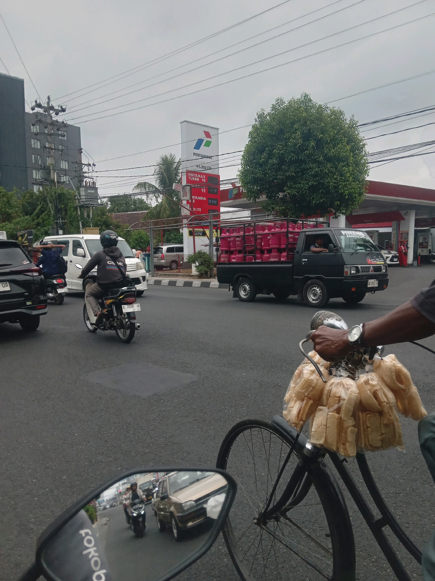 The image depicts a street scene in Depok, Indonesia, characterized by overcast skies and moderate traffic. A black pickup truck, loaded with several pink LPG gas cylinders, is the most prominent vehicle in the mid-ground, driving on the right side of the road. The truck has a license plate that appears to read "AA 8202 IB". To the left of the truck, a white van is also visible.
In the foreground, the handlebars and front wheel of a bicycle are visible, laden with several plastic bags of what appear to be snacks or food items, possibly crackers or puffed rice. The rider's hand, wearing a watch, is visible on the handlebar. A motorcycle with a rider wearing a helmet and a gray hoodie is in the immediate left foreground, its reflection visible in the side mirror of the bicycle. The side mirror shows other vehicles in traffic, including a beige car.
In the background, a Pertamina gas station is clearly identifiable with its red and white signage and fuel pumps. A tall, dark building with multiple windows is situated to the left of the gas station. Overhead power lines crisscross the sky. Trees, including a large leafy one and a palm tree, are present, interspersed with utility poles and traffic infrastructure. The road surface is asphalt, with visible lane markings and some patchy areas. The overall impression is that of a busy urban environment on a cloudy day.