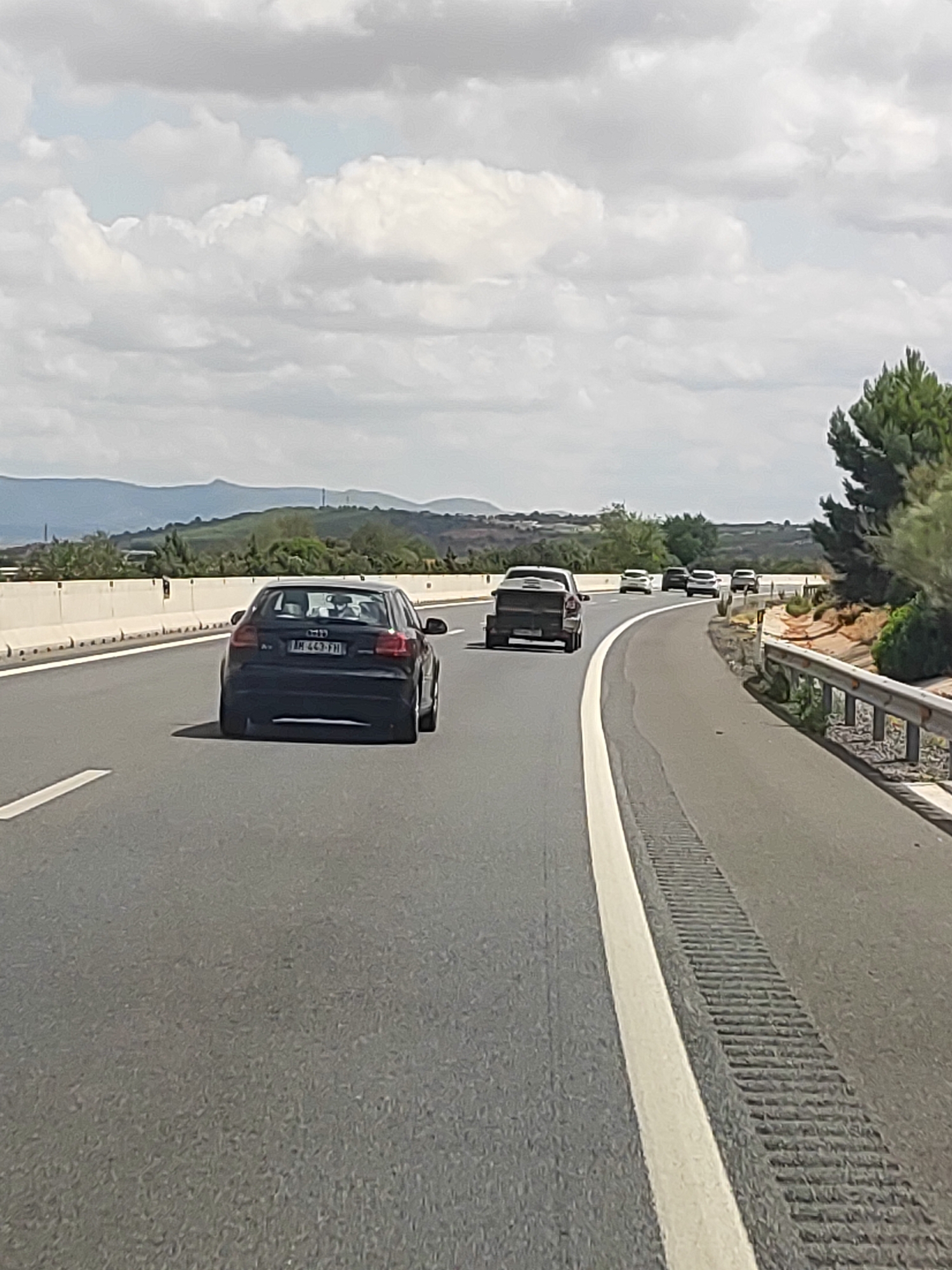 A black Audi A3 is driving on a highway in Fuenterrobles, Spain. The car's license plate is "AH 443 FFI". There are other cars on the highway, including a black pickup truck in front of the Audi and several white cars further down the road. The highway has two lanes in each direction and is surrounded by a natural landscape with hills and trees. The sky is cloudy and the sun is shining. The time of day appears to be daytime, likely afternoon.