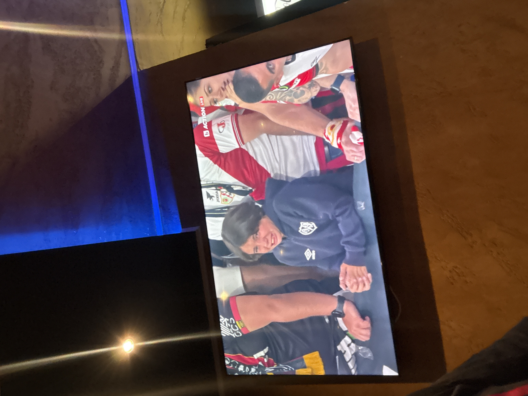 A television screen displays a live sporting event, possibly a fight, with two individuals locked in a grappling hold. A third person, wearing a blue sweatshirt and facing the camera, is smiling. The room appears to be indoors, with a blue-lit wall in the background. There are no immediate indicators pointing towards Burgas, Bulgaria in the image.
