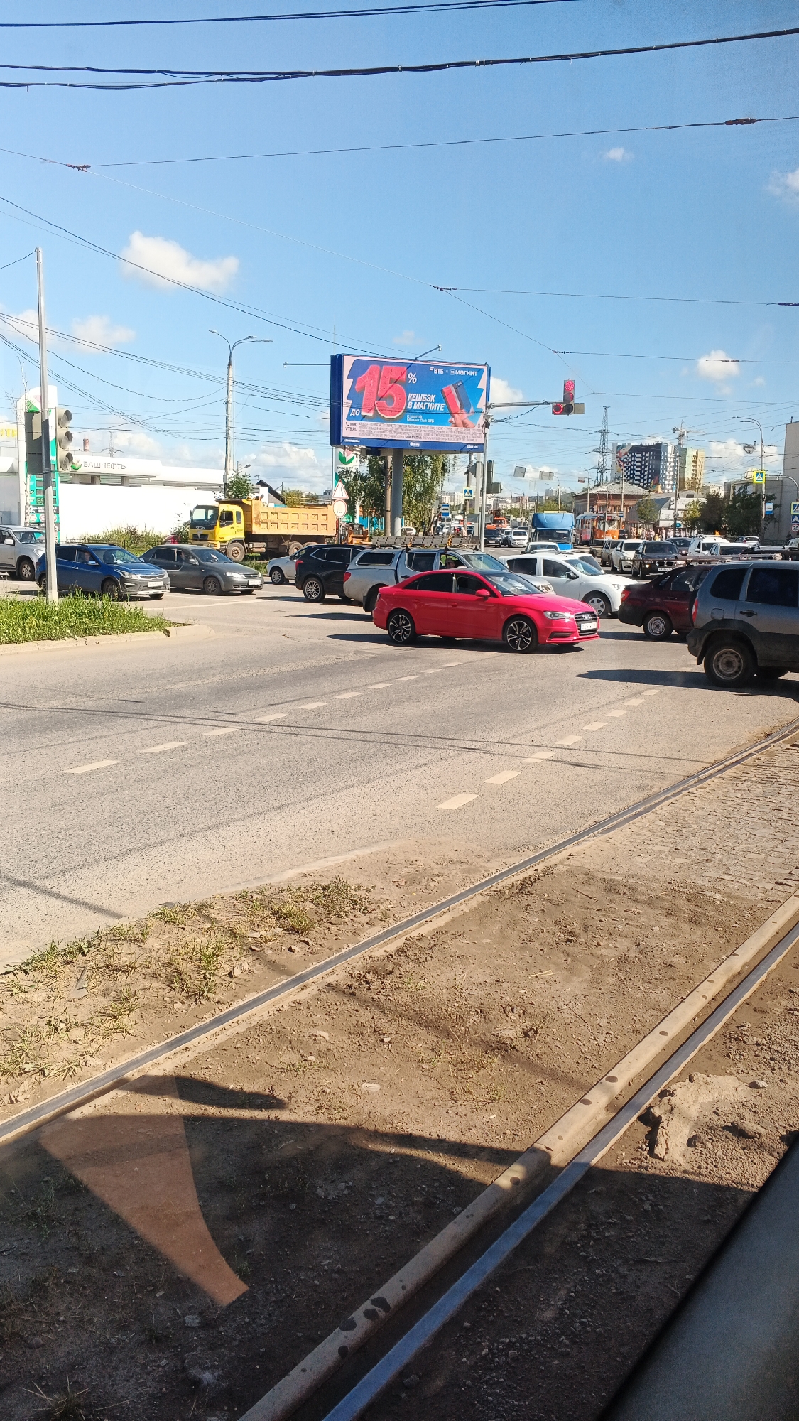 The image depicts a street intersection in Izhevsk, Russia, on a sunny day. In the foreground, trolley tracks run across the asphalt road, with dirt and sparse vegetation on the side. Several cars are present, including a red sedan in the middle of the intersection, a grey sedan to its left, and a black SUV behind it. A yellow truck with a dump bed is parked on the side of the road in the background. Above the intersection, a large billboard displays an advertisement for a promotion, featuring the number "15%" prominently. Traffic lights are visible, with one showing red. Buildings and trees are scattered throughout the urban landscape. The sky is blue with a few scattered clouds.