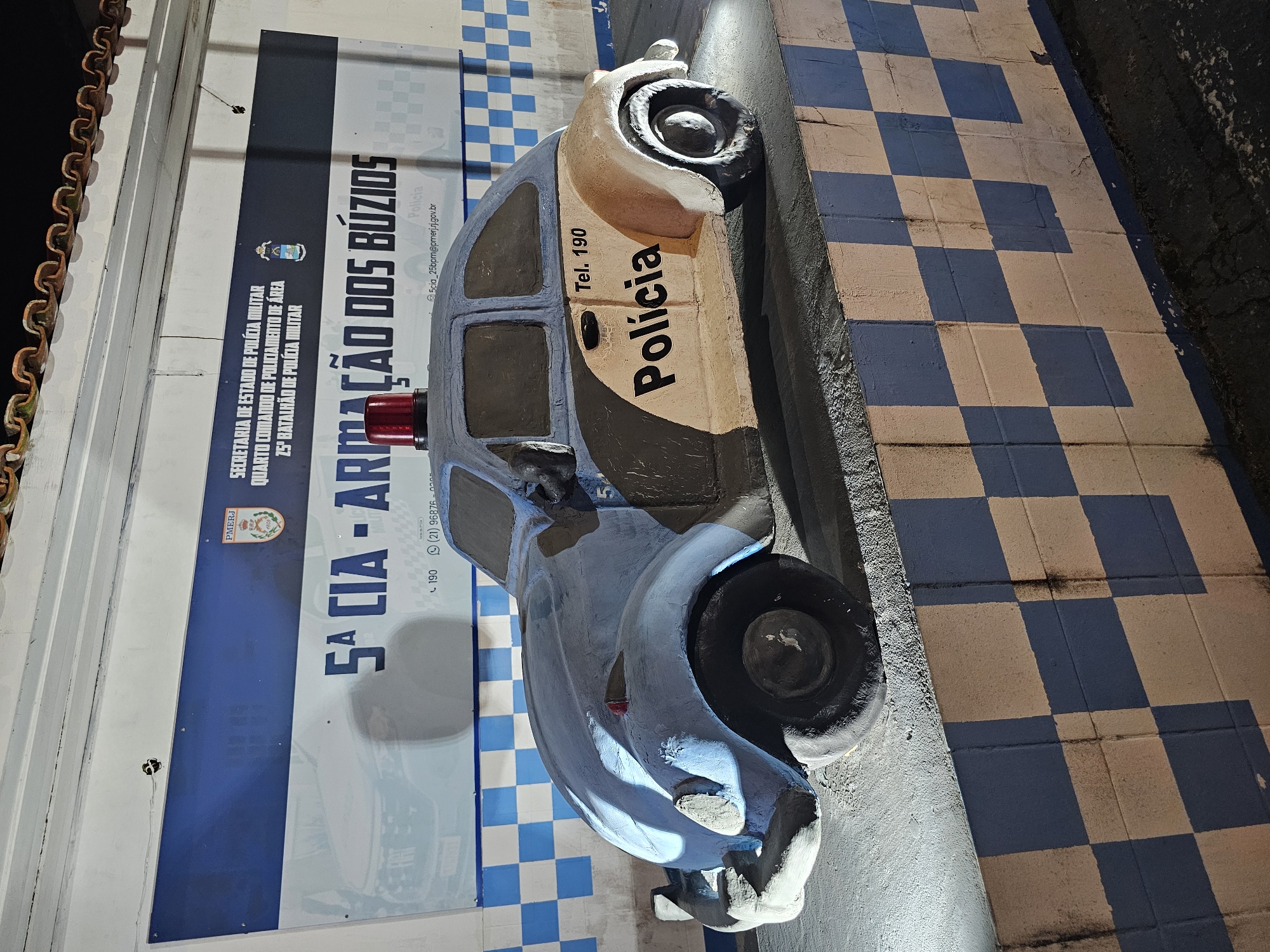 The image shows a concrete sculpture of a police car, painted in a stylized light blue and beige color scheme with black wheels, parked in front of a wall with a blue and white checkered pattern and a sign for the 5th CIA of the 25th Battalion of the Military Police in Armação dos Búzios, Brazil. The sign includes the contact information "Tel. 190" and "5cia_25bpm@pmerj.rj.gov.br". The sculpture sits on a curb with a similar blue and white checkered pattern. The photograph appears to be taken at night or in a dimly lit area.