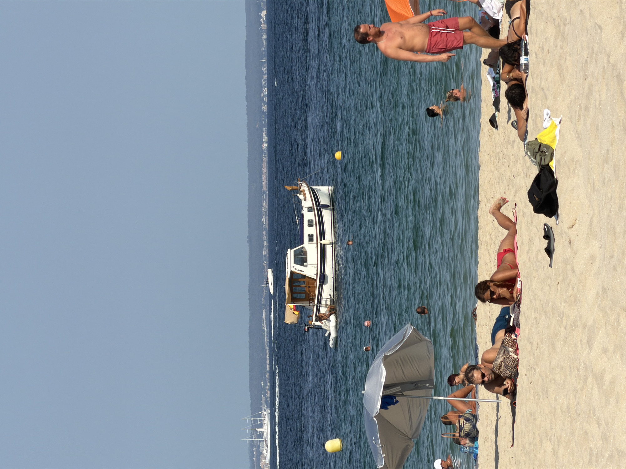 In Palmanova, Spain, a sunny day unfolds on a sandy beach with people enjoying the water and sand. In the foreground, several individuals are relaxing on towels. A man in blue shorts is looking at his phone while lying on the sand, next to him a woman in a red bikini is sunbathing. Further down the beach, another couple is lying on towels, with one person wearing a black bikini. To the right, a man in red shorts is standing and walking, with an orange umbrella partially visible. In the water, several people are swimming and visible from the chest up. A white boat with a brown canopy is anchored in the blue sea, with a smaller inflatable boat tied to its side. A yellow buoy floats in the water near the boat. In the background, a large white sailboat with three masts is visible on the horizon, along with a distant shoreline with buildings. The sky is clear and blue.