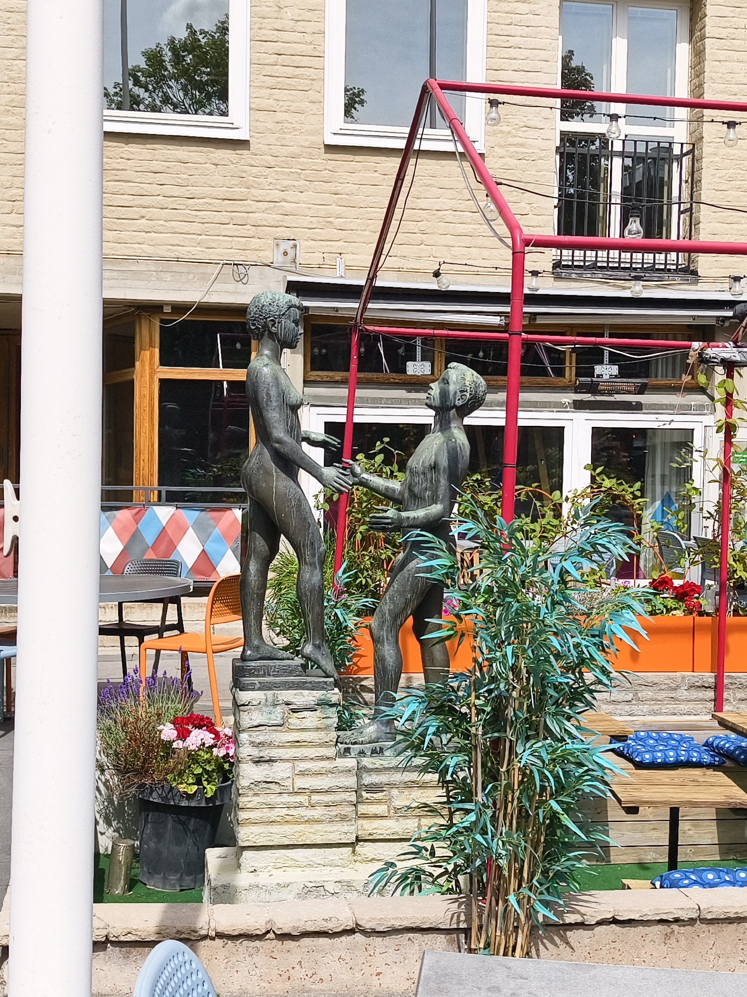 The image depicts an outdoor seating area with two bronze statues in the foreground. The statues are of human figures, one appearing to be female and the other male, standing on a multi-tiered stone pedestal. Both statues are unclothed and appear to be interacting with each other. Behind the statues, there is a building with a light-colored brick facade and large windows. A red metal structure with string lights runs across the scene overhead.
The seating area includes a table and chairs, as well as wooden benches with blue cushions. A large black planter filled with flowers, including lavender and red and pink blossoms, is positioned to the left of the statues. A vibrant green, artificial-looking plant with bamboo-like stalks stands in front of the right statue. The ground is covered in artificial turf.
The location is identified as Linköping, Sweden, specifically the courtyard of the restaurant Frimis Kungshall.