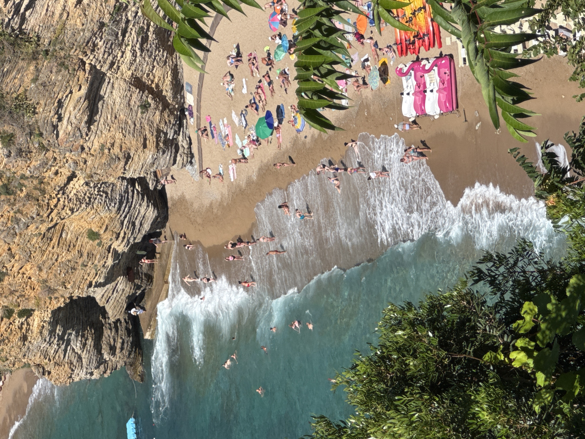 This is an aerial photograph of Mogren Beach in Budva, Montenegro. The beach is characterized by golden sand and is nestled against a steep, rocky cliff face. The turquoise water of the Adriatic Sea laps at the shore, with waves creating white foam. A section of the beach is occupied by numerous sunbathers, many lying on towels or under colorful umbrellas. Several people are wading or swimming in the sea. In the foreground, lush green foliage partially obscures the view. On the sand, there are several pink flamingo-shaped paddle boats and red and yellow kayaks, suggesting recreational activities. The cliff face features layered rock formations, with some sparse vegetation growing on it. A small concrete platform extends into the sea from the base of the cliff.