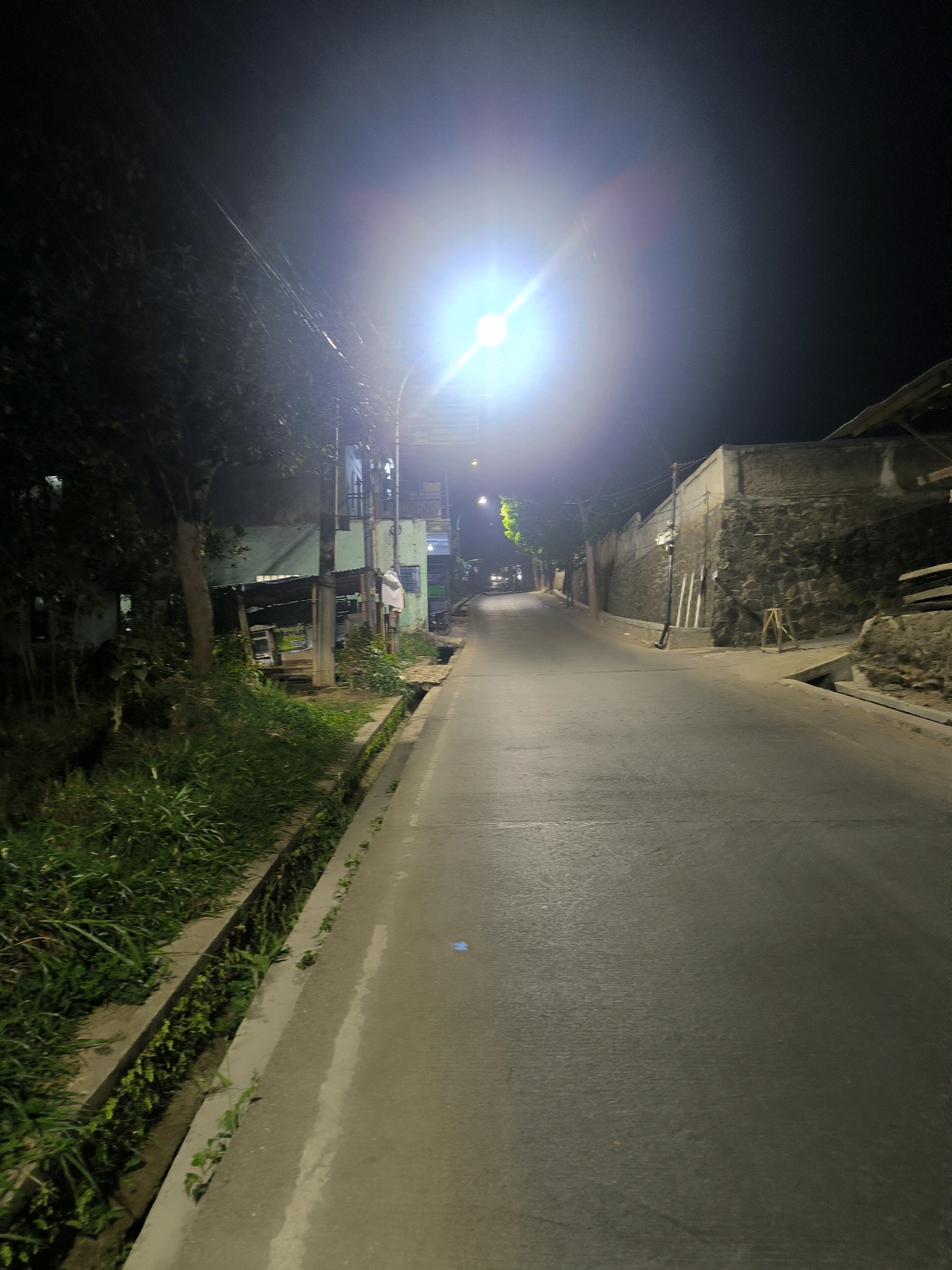 The image depicts a street scene at night, illuminated by a bright streetlamp. The road is paved and curves to the right, receding into the darkness. On the left side of the road, there is a drainage ditch filled with vegetation, followed by dense greenery and trees. Beyond the trees, some buildings are visible, including a two-story structure with balconies and a green facade. On the right side of the road, a low stone wall borders the street, with some construction materials or debris piled nearby. The sky is dark, with the strong light from the streetlamp causing lens flare. The scene appears to be in a residential or semi-urban area, likely in Bandung City, Indonesia, given the typical infrastructure and vegetation seen. There are no people or vehicles clearly visible. The overall impression is a quiet, somewhat unkempt street at night.