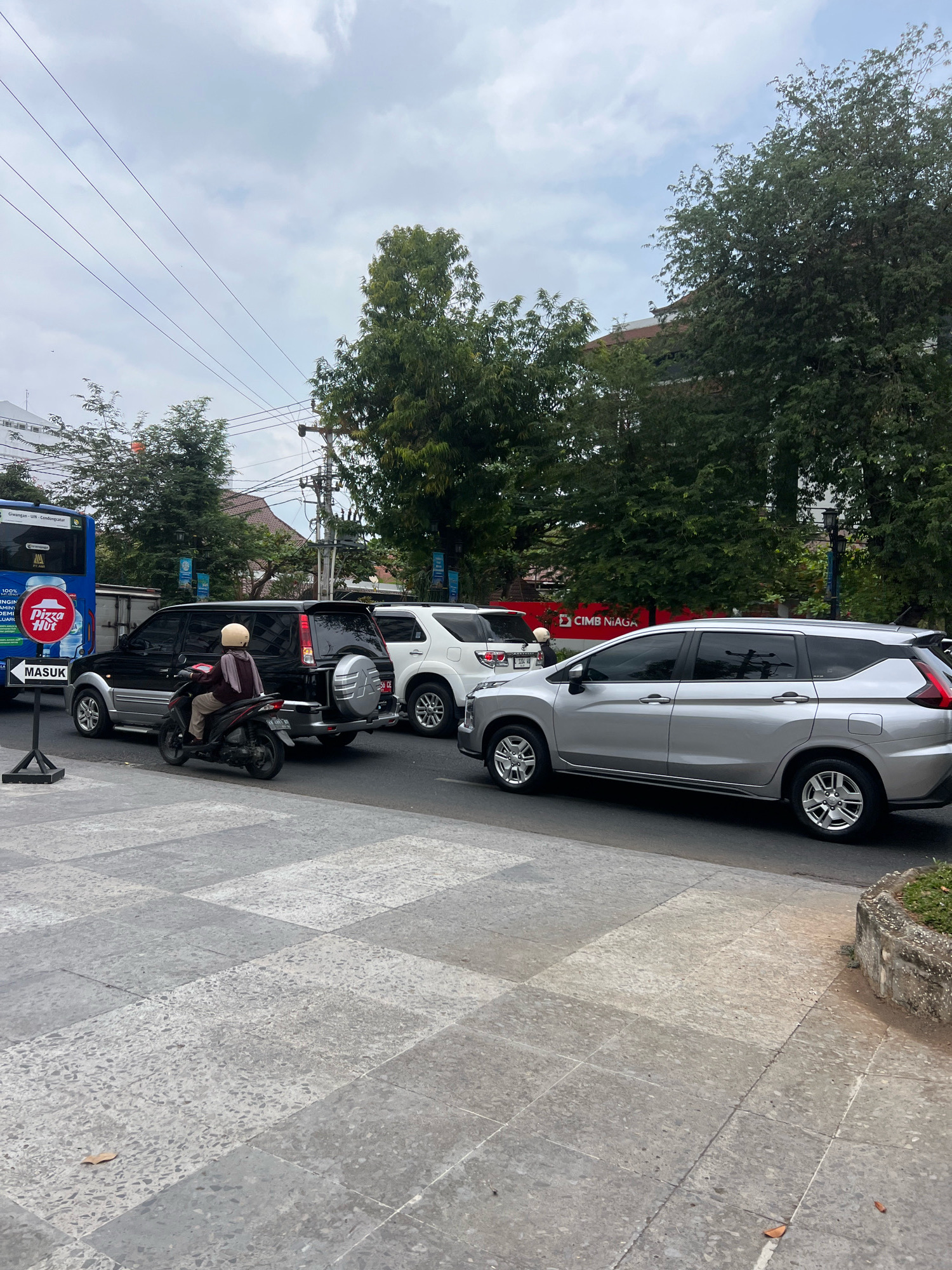 The image depicts a street scene in Yogyakarta, Indonesia, on a cloudy day. In the foreground, a motorcycle with a rider wearing a light-colored helmet and a dark maroon jacket is stopped in traffic. The motorcycle is a dark color with red accents. Behind the motorcycle is a black SUV with a spare tire mounted on the rear. To the right of the black SUV is a white SUV. Further to the right is a silver MPV. A blue bus is visible on the far left of the image. A red sign with a white "Pizza Hut" logo and the word "MASUK" (Indonesian for "enter") in white text on an arrow pointing left is positioned on a black stand in the lower left portion of the frame. The street is paved, and there is a sidewalk made of large, light-colored square tiles. Trees line the background, and a building with a red awning is visible behind the trees. Utility poles and wires are also present in the background. The overall impression is a typical urban traffic scene.