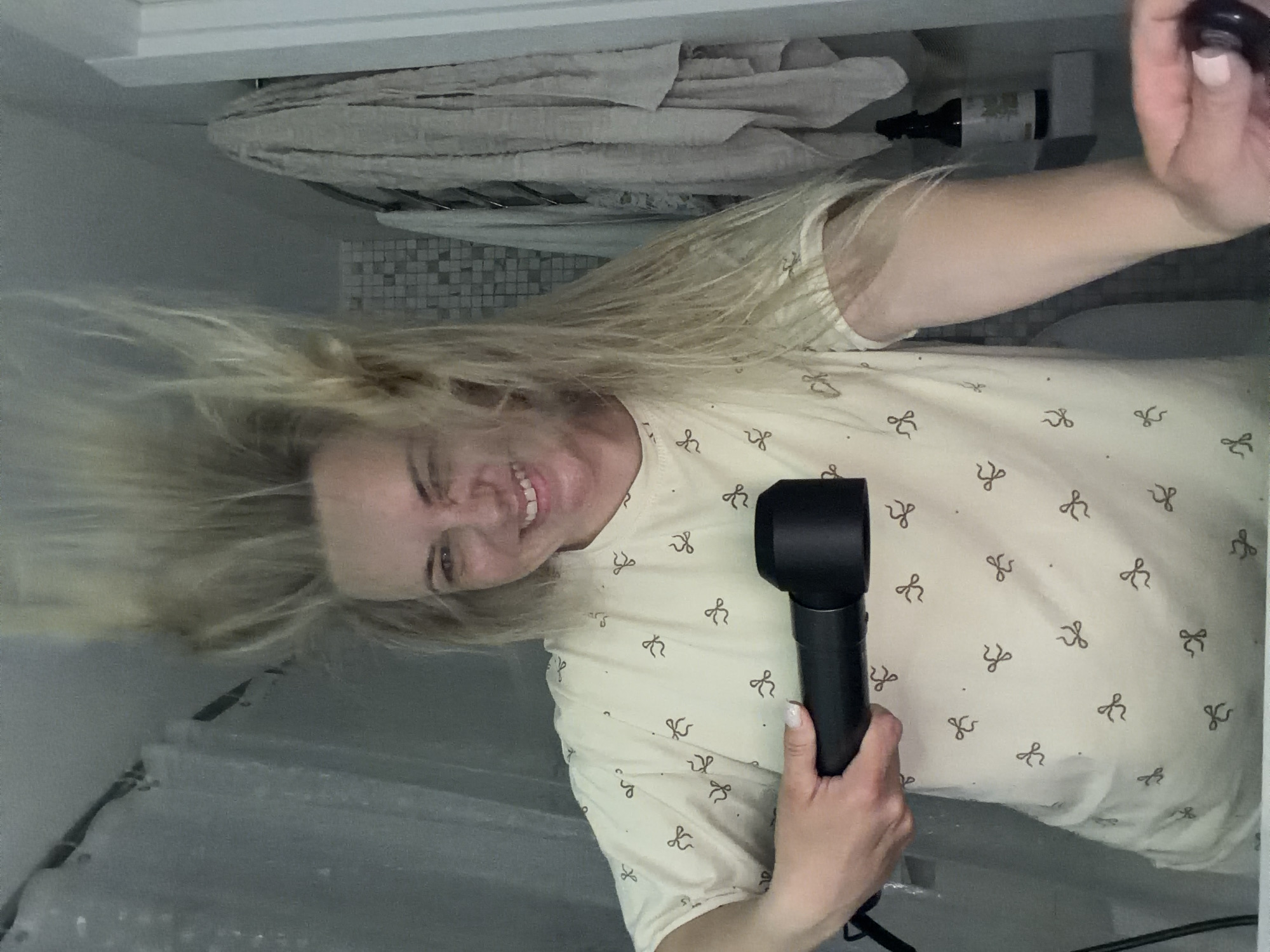 The image displays a blonde woman actively drying her hair in what appears to be a bathroom. Her hair is dynamically fanned out and flying upwards, indicating the force of the black, modern hair dryer she holds in her right hand. She is smiling brightly and looking towards the camera, suggesting a self-portrait. She wears a light-colored t-shirt featuring a repeating pattern of small, dark bow-tie or ribbon shapes. Her fingernails are painted a light pink.
The background details support the bathroom setting. To her upper left, folded light-colored towels are visible on a shelf or rack, with a dark bottle positioned nearby. Below these, there is a section of wall with small, possibly mosaic, tiles. A textured, translucent surface, likely a shower curtain or screen, is visible in the lower part of the frame. The lighting is bright and even, typical of an indoor environment. The woman's expression conveys happiness or playfulness. No meaningful or legible text is visible within the image. This scene is set in Votkinsk, Russia.