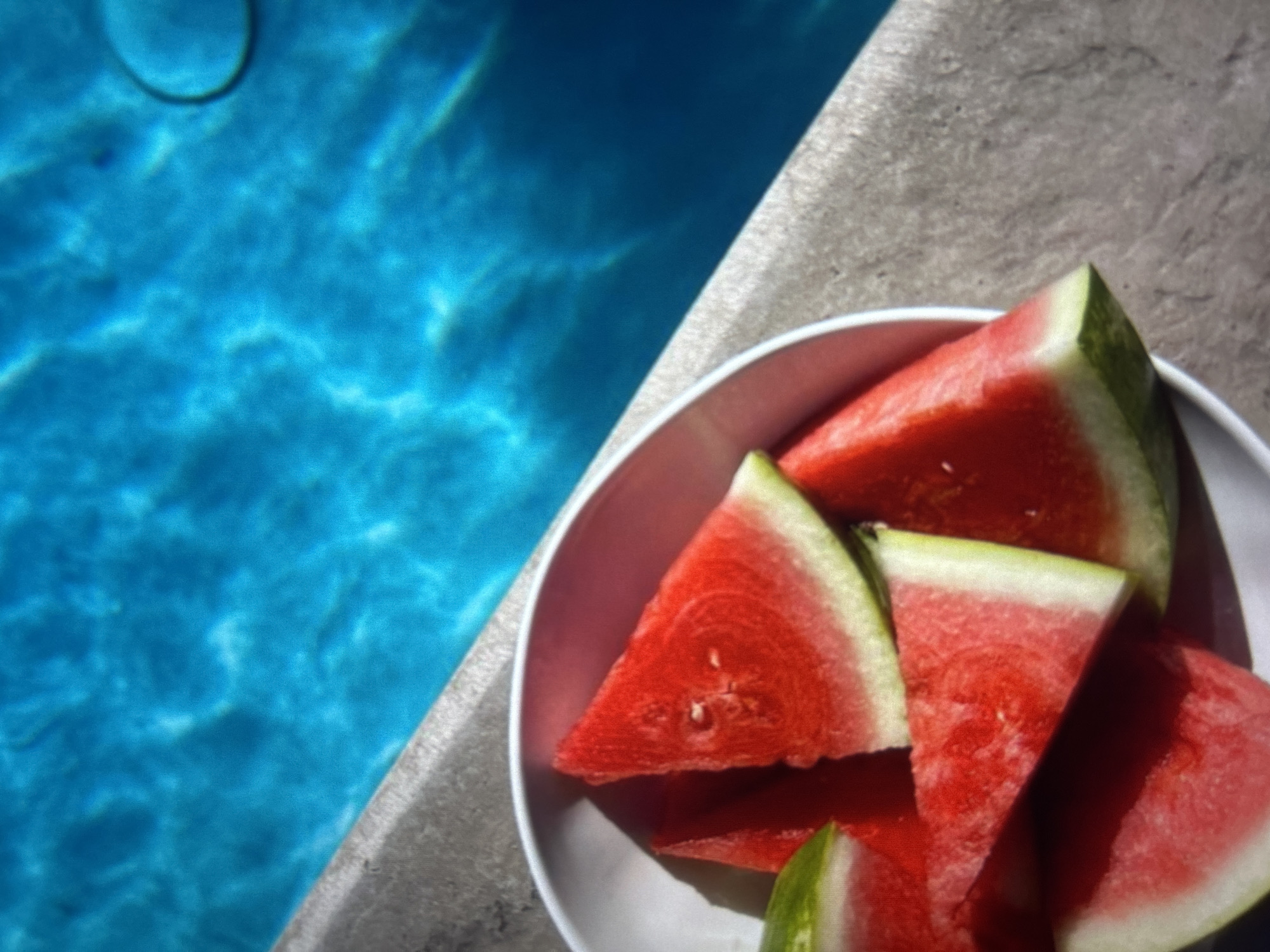 The image shows slices of watermelon in a white bowl, positioned near the edge of a swimming pool. The pool water is a bright blue, with sunlight reflecting off its surface, creating patterns of light and shadow. The edge of the pool is a textured, grey concrete material. The scene is outdoors, suggesting a warm, sunny day. There is no indication of the city of Knezha, Bulgaria, in the image.