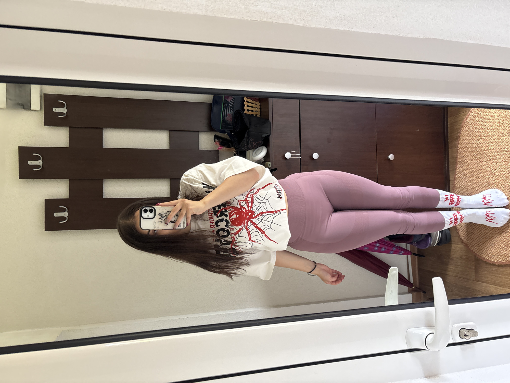 A female is reflected in a mirror, taking a photo with her phone. She wears a white t-shirt with a red spider graphic, pink leggings, and white socks with red lettering. In the background, there is a dark brown wall-mounted coat rack with metal hooks, a closed cabinet above the coat rack, an open bag, shoes, an umbrella, and a circular rug. The mirror is surrounded by a white frame with a white handle. There is no information to indicate this scene is located in the city of Polena, Bulgaria.