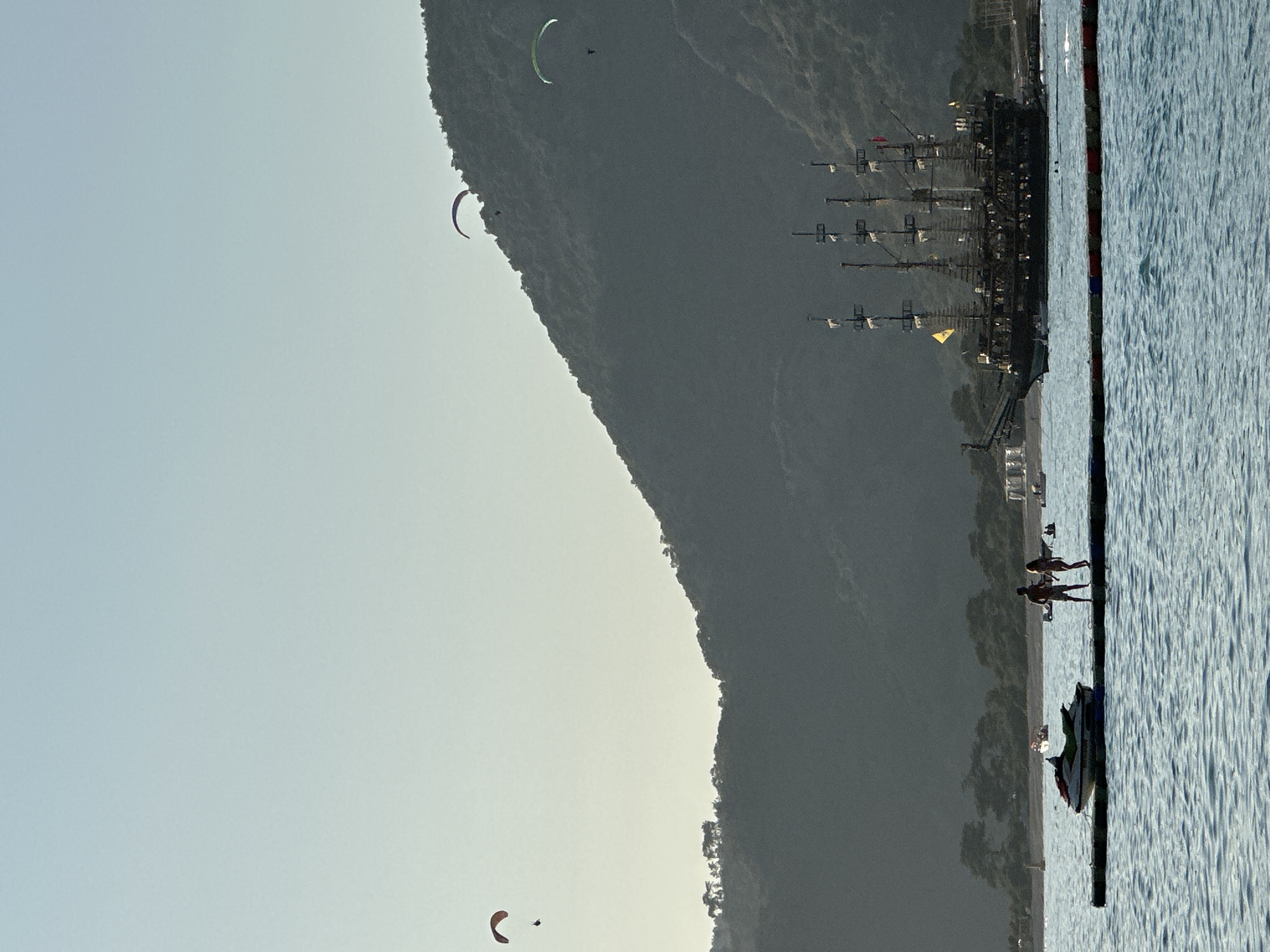 This image captures a vibrant coastal scene in Fethiye, Turkey, featuring a mix of natural beauty and recreational activities. The dominant feature is a large, dark, tree-covered mountain or hill that slopes down from the right side of the frame, meeting a calm body of water on the left.
In the sky above and around the mountain, several paragliders are visible. One prominent paraglider with a bright green canopy is seen high above the mountain's peak, while two others with darker, possibly reddish-brown canopies are lower, closer to the water. The sky itself is a clear, pale blue-grey, suggesting fair weather.
At the base of the mountain, resting in the water, is a large, multi-masted ship, reminiscent of a pirate vessel, with its dark hull and rigging sharply silhouetted. Further towards the foreground, on a floating pier or platform in the water, two people are standing, silhouetted against the bright water. Nearby, a jet ski is partially visible.
The time of day appears to be either late afternoon or early morning, given the soft, ambient light and the shadowed side of the mountain against the brighter sky. The scene conveys a sense of outdoor leisure and adventure in a picturesque mountainous and maritime setting. No discernible text is visible in the image.