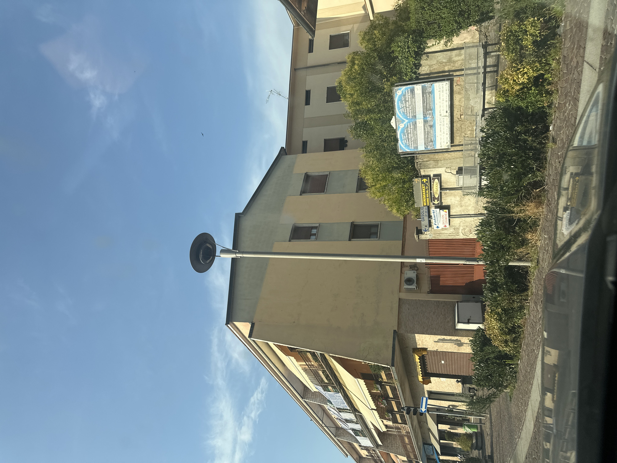 The image shows a street scene in Rudiano, Italy. The focus is on a multi-story building with a light yellow facade and balconies with awnings. A tall lamp post stands in front of the building. To the right of the building, there is a large billboard with Italian text, possibly an advertisement or event poster. Green bushes and a paved area with a cobblestone pattern are visible in the foreground, with the reflection of a car's dashboard on the lower part of the image. The sky is clear blue with a few wispy clouds. The overall impression is a sunny day in a European town.