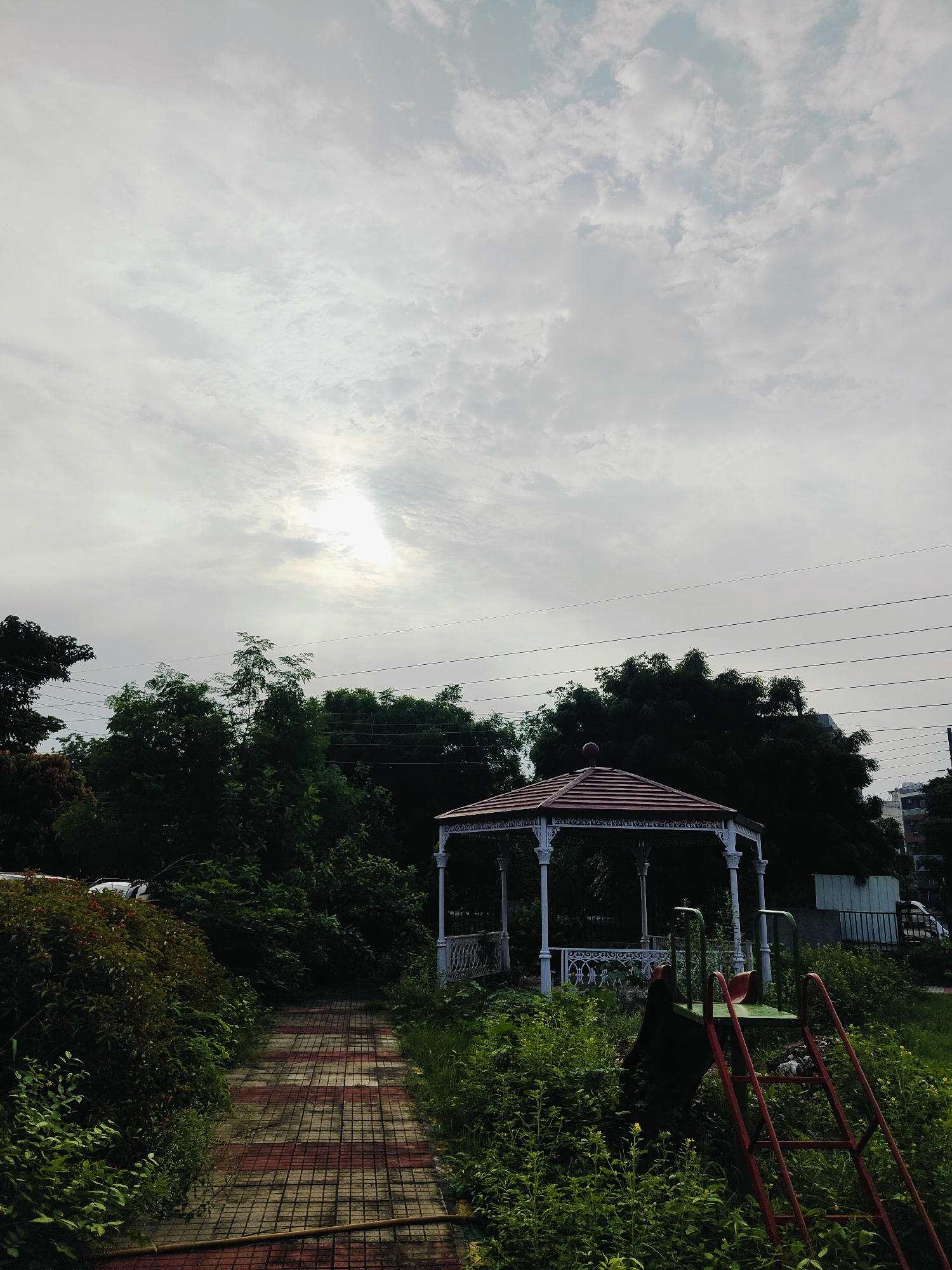 The image depicts a slightly overcast day in Gurgaon, India. In the foreground, a red and white checkered pathway leads into a park area. To the left of the path is dense, green foliage and bushes. To the right, a red metal slide with green steps stands amidst overgrown grass and weeds. Further into the park, a white gazebo with a red roof is visible. Behind the gazebo, a line of dark green trees forms a backdrop, with power lines strung across them. The sky is predominantly cloudy with a patch of bright sunlight breaking through. In the far background, several buildings can be seen.