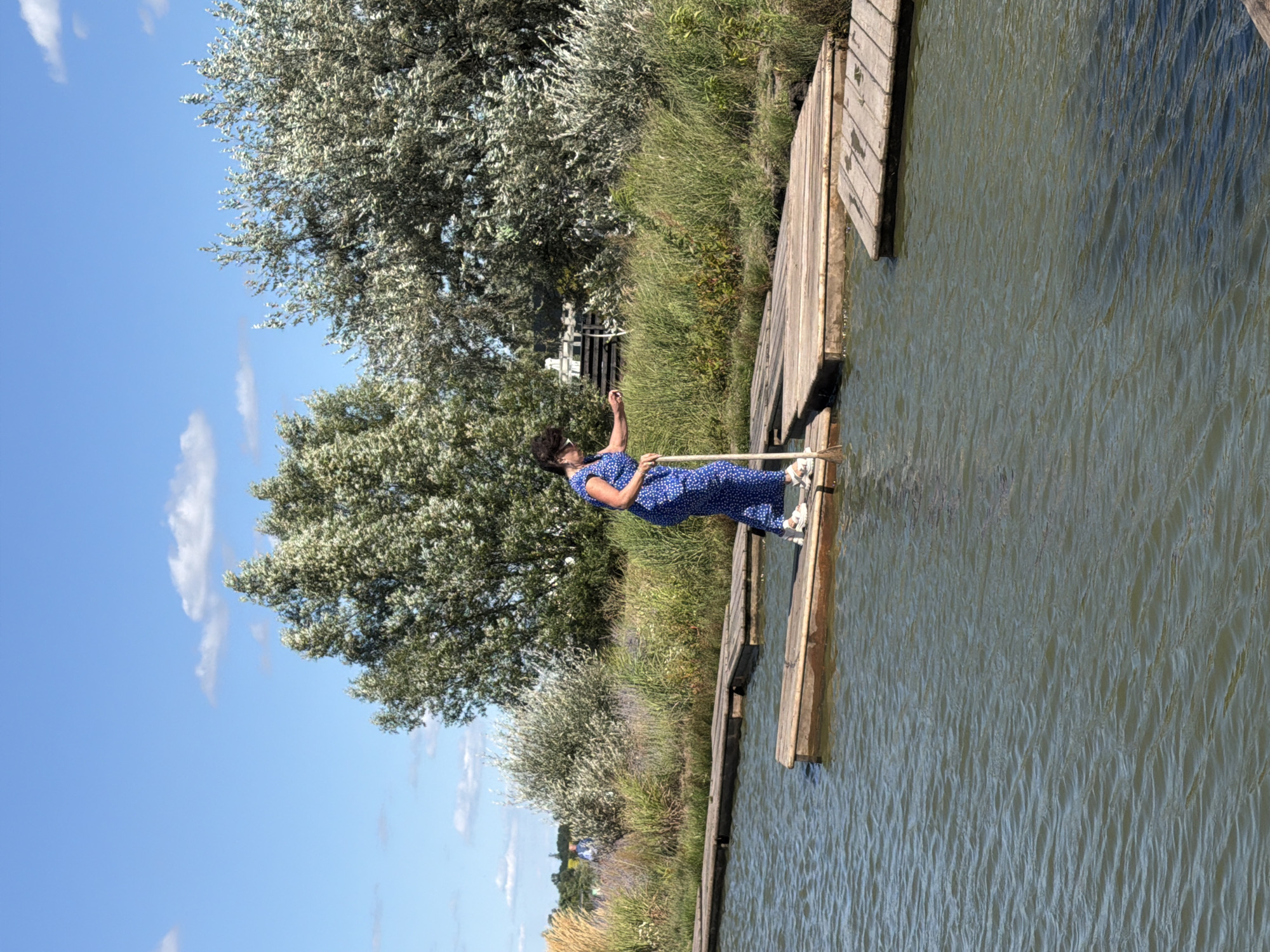 In Severynivka, Ukraine, a woman in a blue polka-dot jumpsuit is walking across a series of wooden planks that bridge a body of water. She is holding a long pole, which she appears to be using for balance. The water is calm with ripples. Tall green grasses and shrubs line the far bank, with lush trees providing a backdrop under a bright blue sky with scattered white clouds.