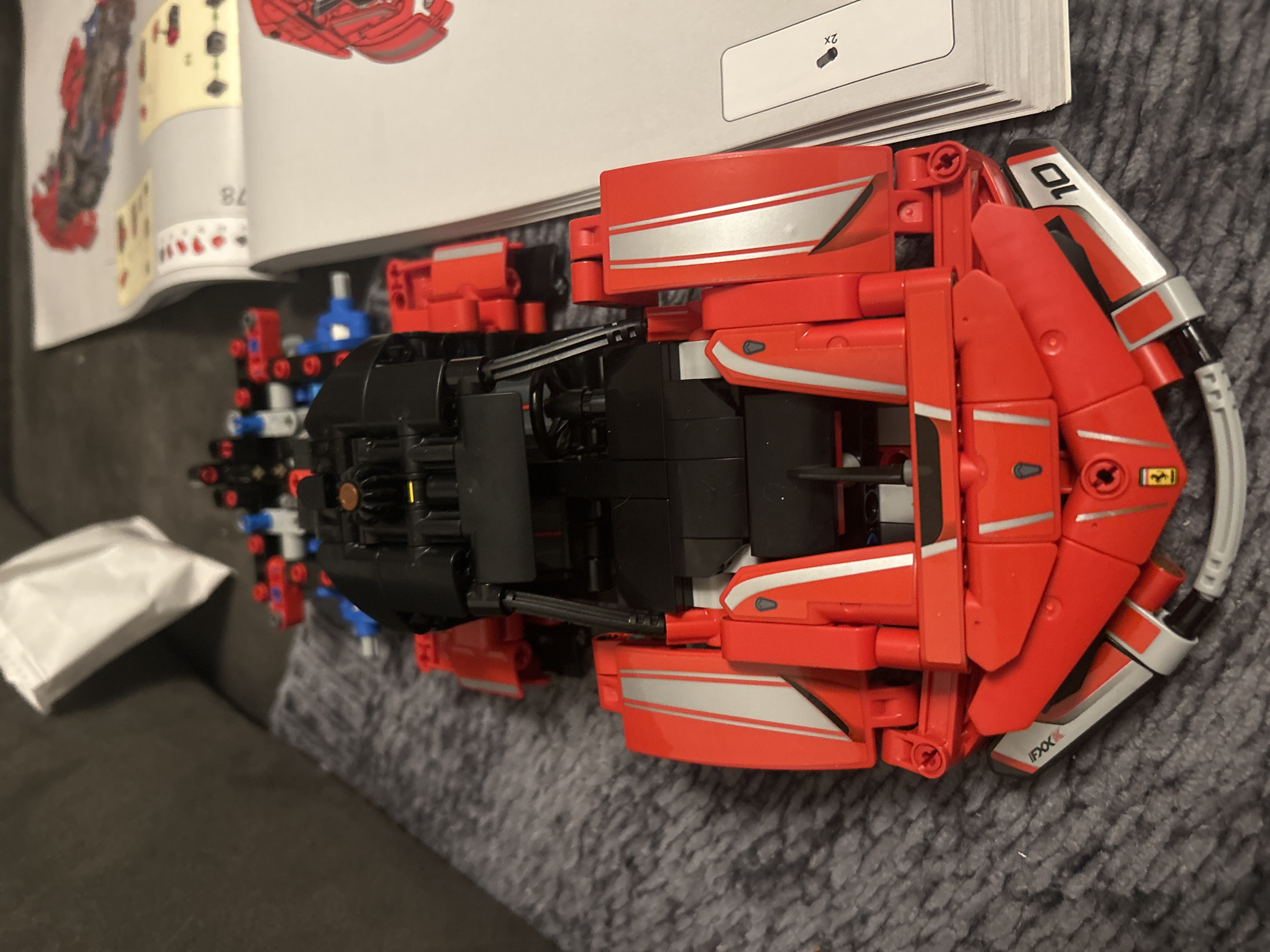 An overhead shot shows a partially assembled red and black LEGO Technic car, likely a model of a Ferrari, resting on a grey textured surface. The car is positioned vertically in the frame, with its front at the bottom. The car features prominent red body panels with silver racing stripes, black aerodynamic elements, and visible LEGO Technic components like gears and axles. Next to the car, to the right, are two open instruction manuals for the LEGO set, with diagrams and page numbers visible. A white paper bag is visible in the upper left corner of the frame, partially obscuring the grey textured surface, which appears to be a couch or rug. The scene is devoid of any human presence or identifiable urban or natural elements within Leiden, Netherlands.