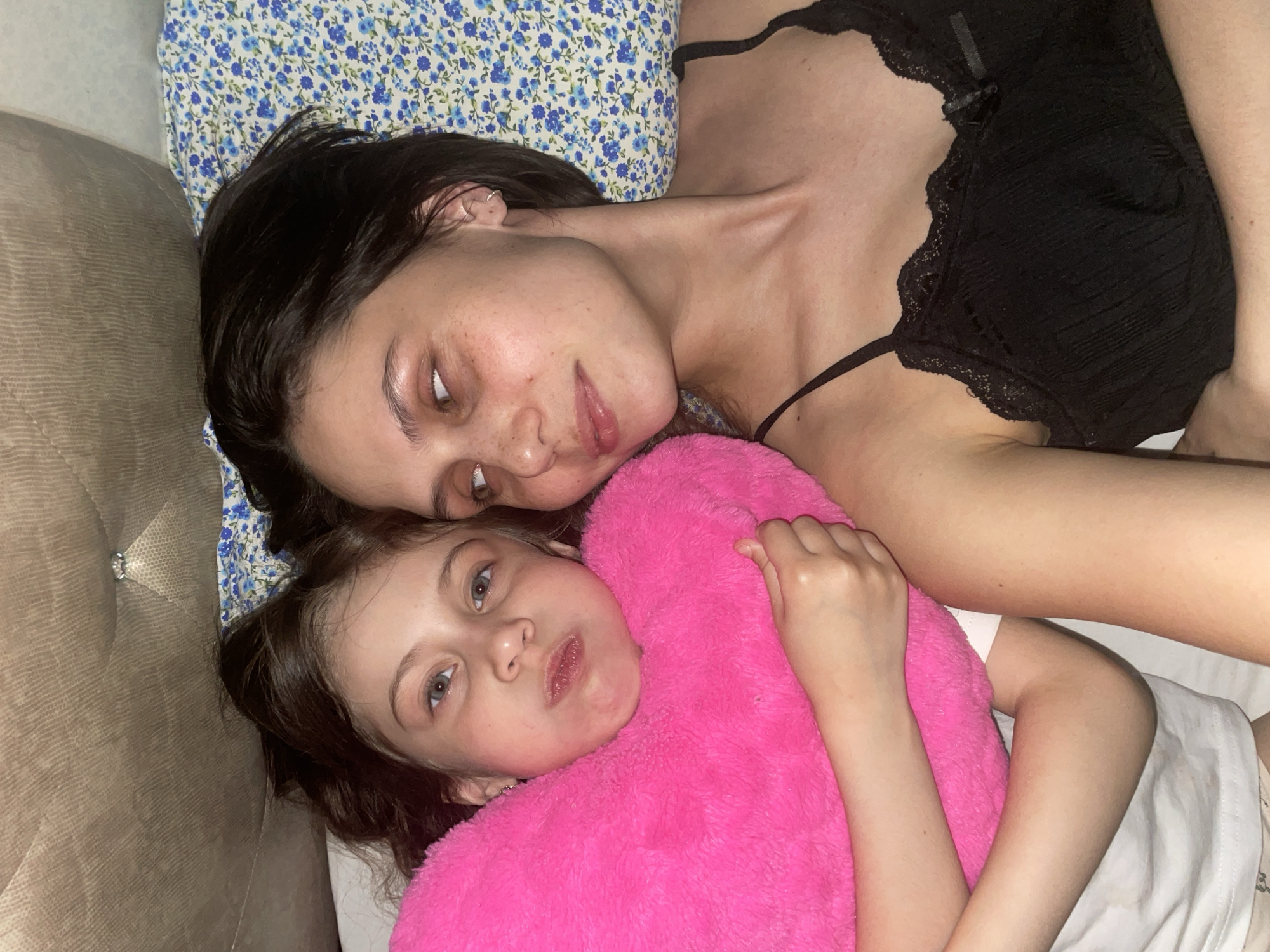 This indoor photograph captures two females, likely a mother and daughter, lying closely together on what appears to be a bed or couch, possibly in a bedroom or living room in Izhevsk, Russia.
The older female, situated towards the top of the frame, has dark hair and a light complexion with visible freckles. She is wearing a black, lace-trimmed camisole and rests her head on a pillow adorned with a blue and white floral pattern. Her eyes are partially closed or softly focused, giving her a serene and relaxed expression as she looks towards the camera.
Beside her, the younger female, a child with light brown hair and blue eyes, is partially covered by a vibrant, fluffy pink blanket or plush toy. She looks directly at the camera with a slightly serious or pouty expression.
The setting appears comfortable and intimate, with soft, ambient lighting suggesting an evening or nighttime scene. A light brown, possibly tufted, piece of furniture serves as a backdrop element, and white bedding is visible beneath the child. The overall mood is one of quiet togetherness. No discernible text is visible in the image.