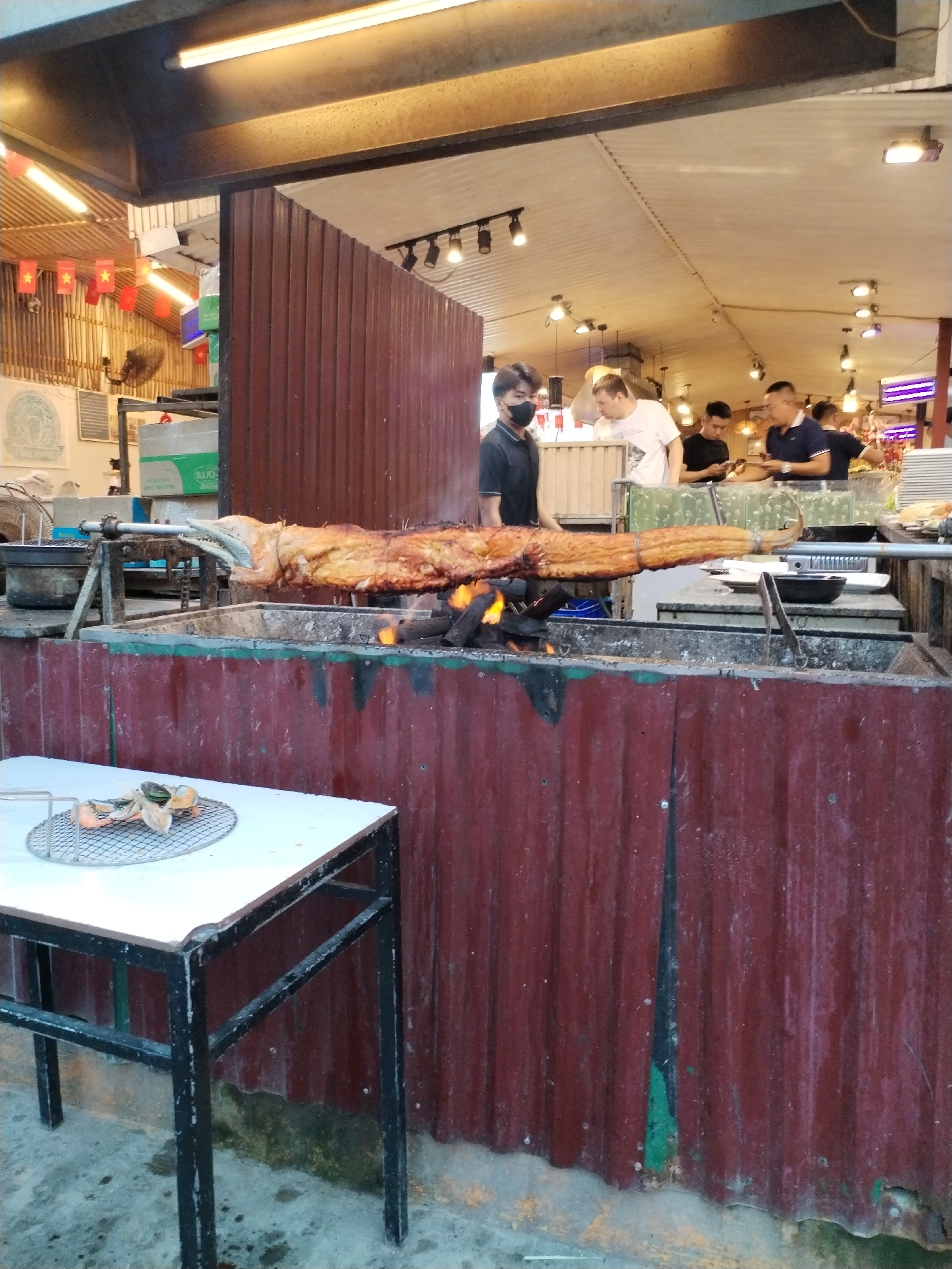 A full alligator is being roasted over an open fire in Bac Nha Trang Ward, Vietnam. The alligator is on a spit, and the fire is directly beneath it. A man wearing a mask stands next to the grill. In the background, other people are standing around. In the foreground, there is a metal table with a wire rack on top, holding cooked food. The grilling area is made of corrugated red metal.