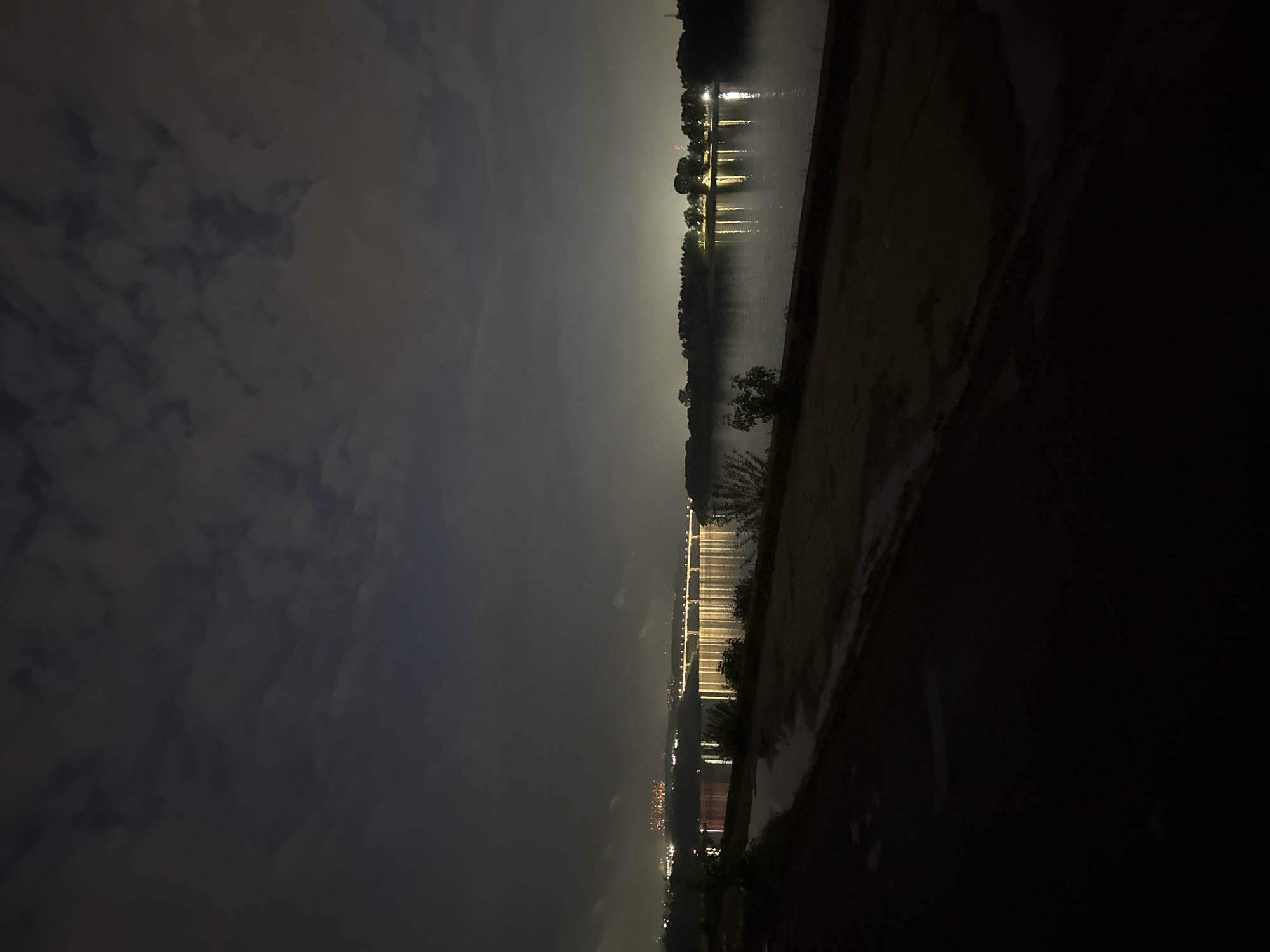 The image shows a nighttime view of a river in Nizhny Novgorod, Russia. A bridge, illuminated with lights, stretches across the water. Reflections of lights are visible on the water's surface. On either side of the river are dark banks with some vegetation. The sky is cloudy. The image appears to be taken from an elevated position.