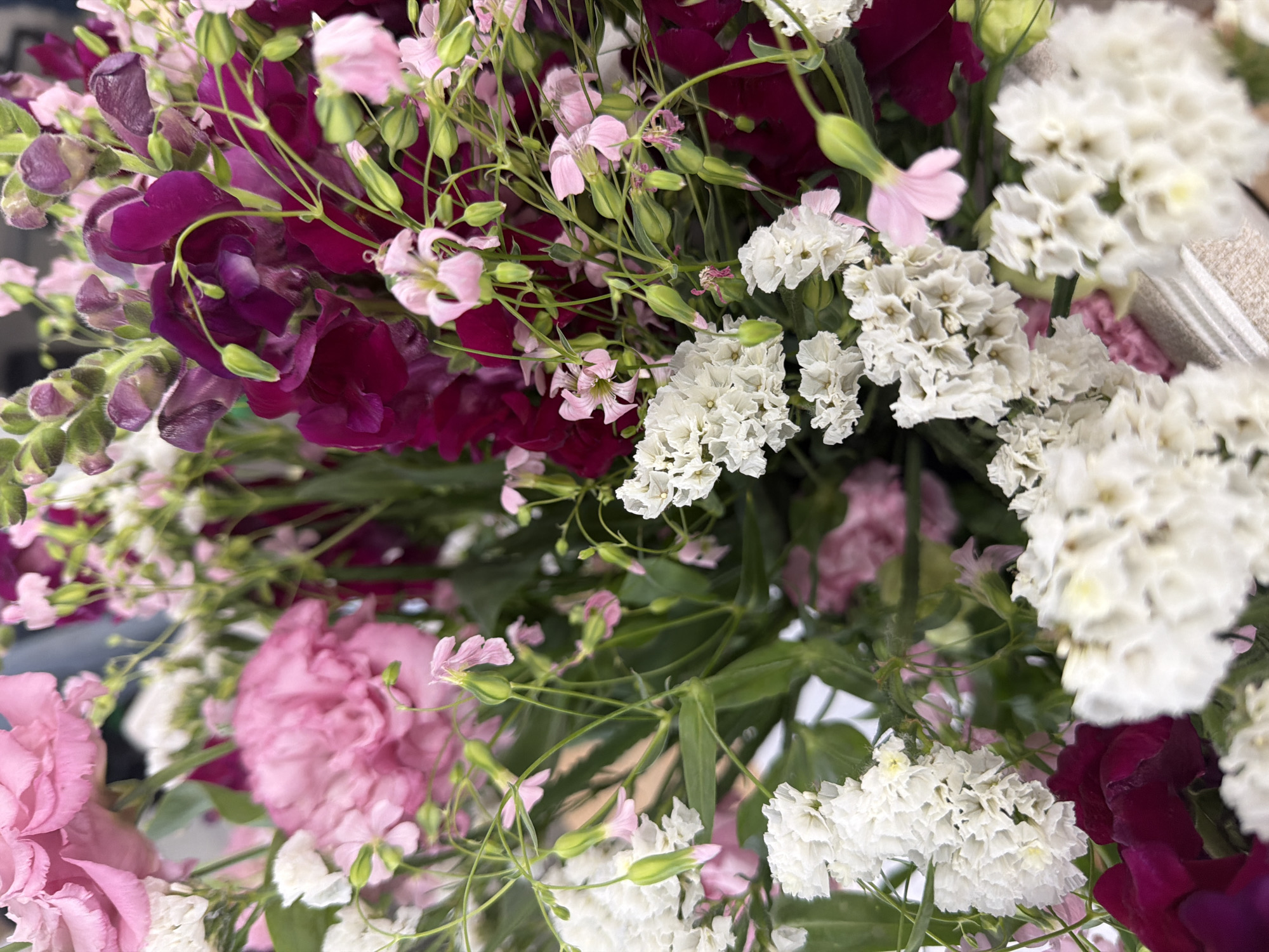 The image shows a bouquet of flowers with various colors and types. There are pink carnations, white statice flowers, magenta sweet peas, and small pink flowers with green stems. The arrangement is densely packed, and the lighting is soft. The location is not discernible from the image.