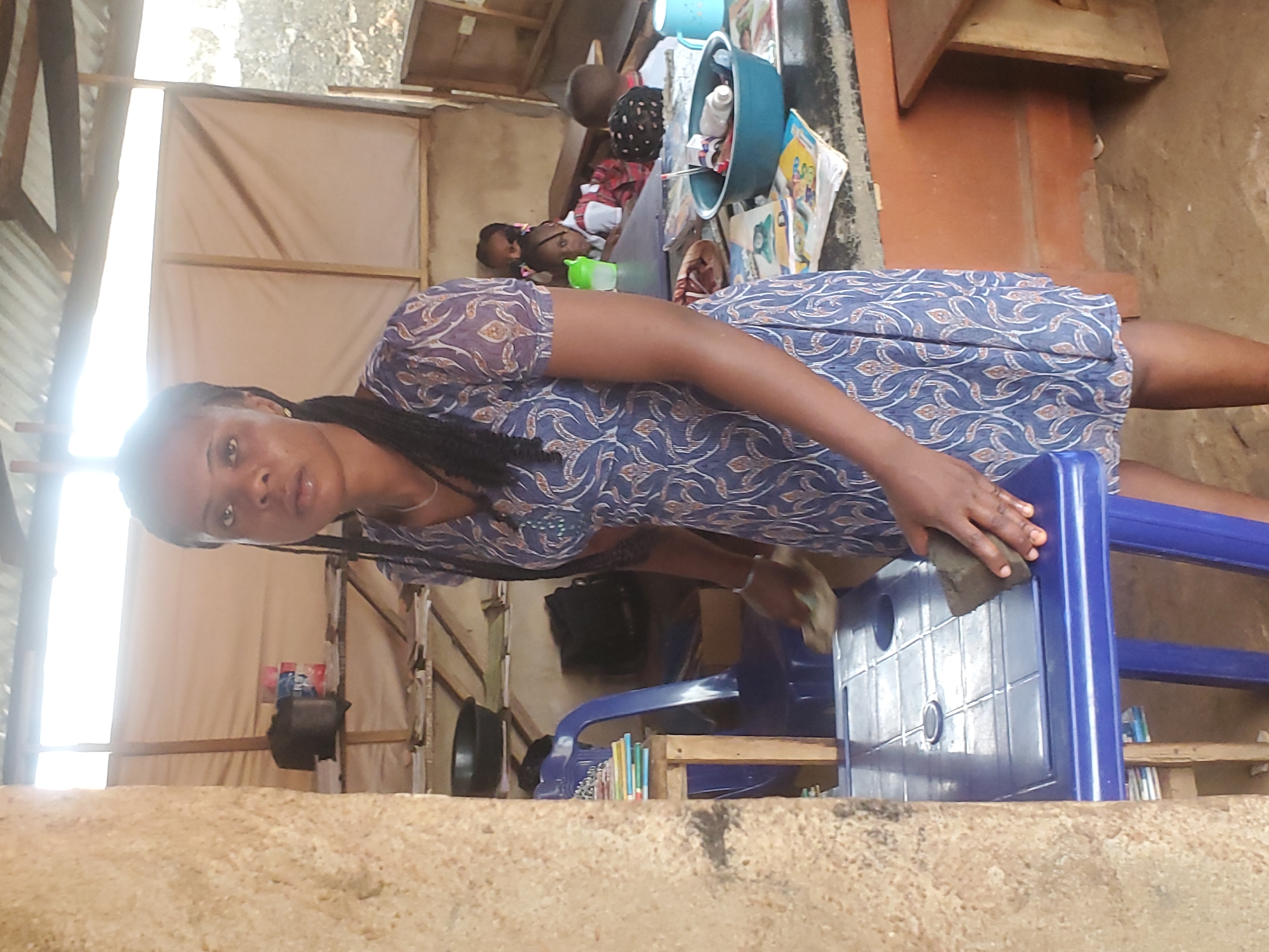 A woman is cleaning a blue plastic chair with a sponge in Ukwu-Abwa, Nigeria. Several children are lying down on a mattress with their backs facing the camera, and a green bowl is next to them.