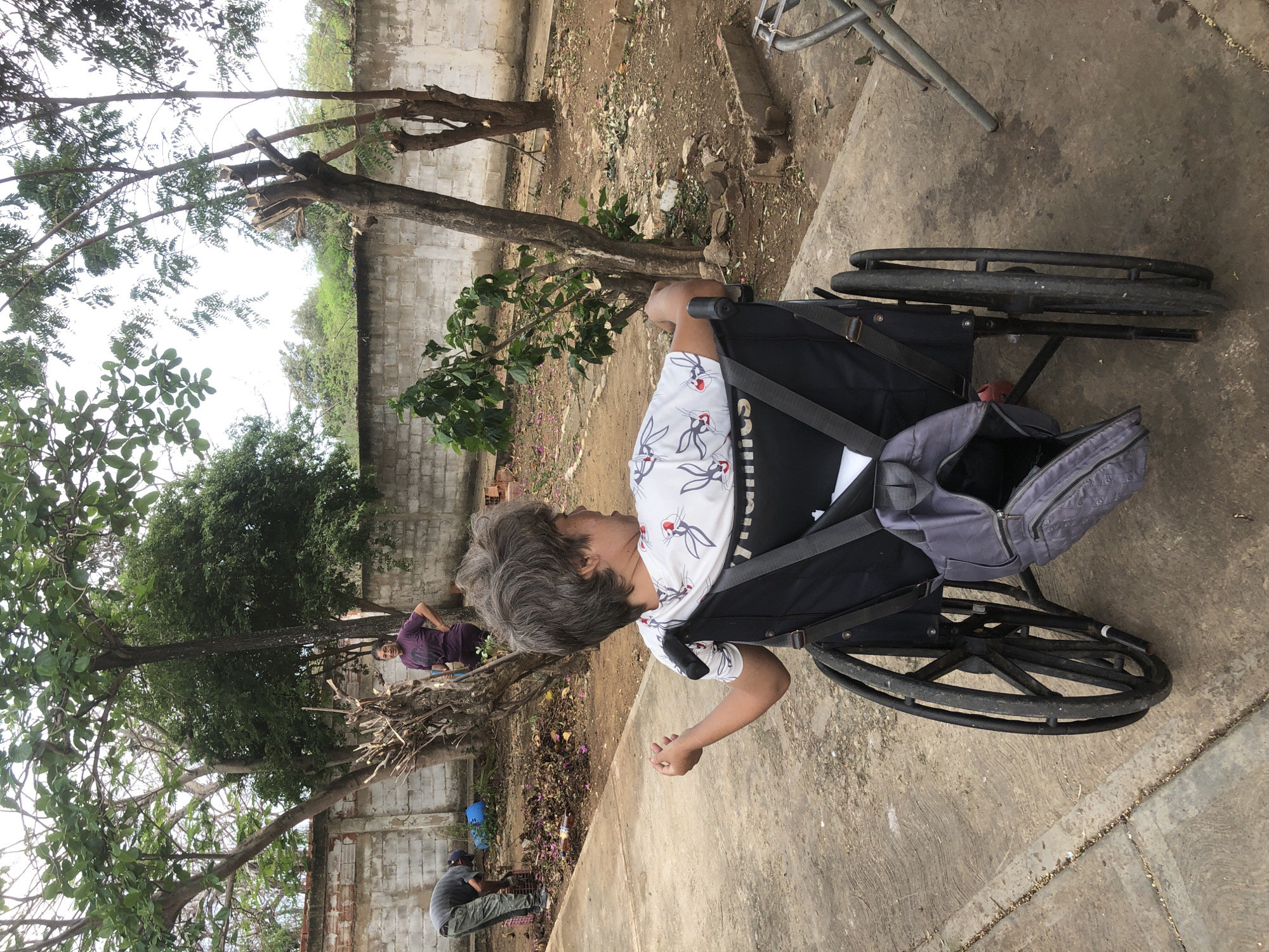 The image depicts an outdoor scene in what appears to be a backyard or communal area in La Tagua, Venezuela. In the foreground, a person is seated in a black wheelchair, viewed from behind and the right side. The individual has gray hair and is wearing a white t-shirt with a repeating pattern of cartoon characters. The wheelchair has a gray bag attached to its back.
In the middle ground, a woman in a purple t-shirt stands near a pile of cut branches and debris. To her left, another person, wearing a gray shirt and pants, is bending over, raking leaves or debris into a crate. A blue bucket is visible near the raking person.
The environment consists of a concrete pathway in the foreground, leading to a dirt and gravel ground area. There are several trees in various states of growth and pruning, with dense foliage visible in the background. A white brick wall runs along the perimeter of the area, partially obscured by trees and vegetation. The overall impression is that of a residential or semi-public outdoor space where yard work is being conducted.