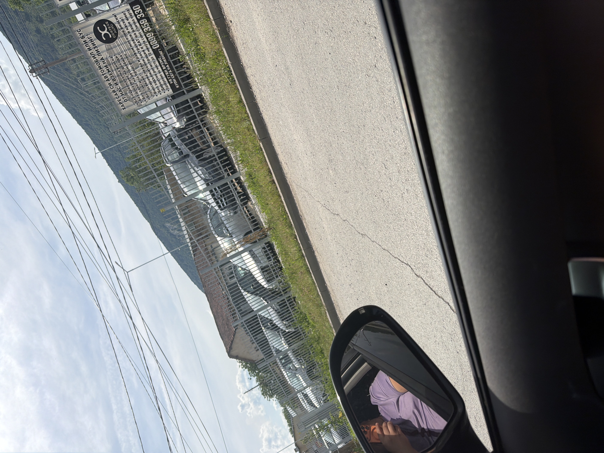 The image, taken from inside a car, shows a bright daytime scene looking out the passenger side window and mirror. On the right, the dark interior of the car's door frame is visible, with a side mirror reflecting a person wearing a purple top, holding what appears to be a phone or small object.
Outside the car, an asphalt road runs parallel to a grassy verge. Beyond the verge, a metal fence encloses what looks like a car dealership or lot, where several white or light-colored cars are parked. In the background, green hills or mountains rise under a sky with scattered white clouds. Utility wires crisscross the upper portion of the sky. Some buildings are partially visible behind the car lot.
A large sign on the fence, written in Cyrillic, advertises car leasing services. The text reads: "Choose a car or van from us and take it on lease!" It lists various terms, including "No initial down payment!", "No income proof!", "No car collateral!", "No early repayment fees!", "Fixed interest and equal monthly installments!", "No mandatory insurance and casco!", "Direct registration at the Traffic Police in your name!", and "Approval on the spot in 10 minutes with just an ID card!" The sign also displays a company name, "CORTEGE AUTO," and a phone number: "0899 359 330." Given the Cyrillic text and provided context, the scene is set in Shumen, Bulgaria.
