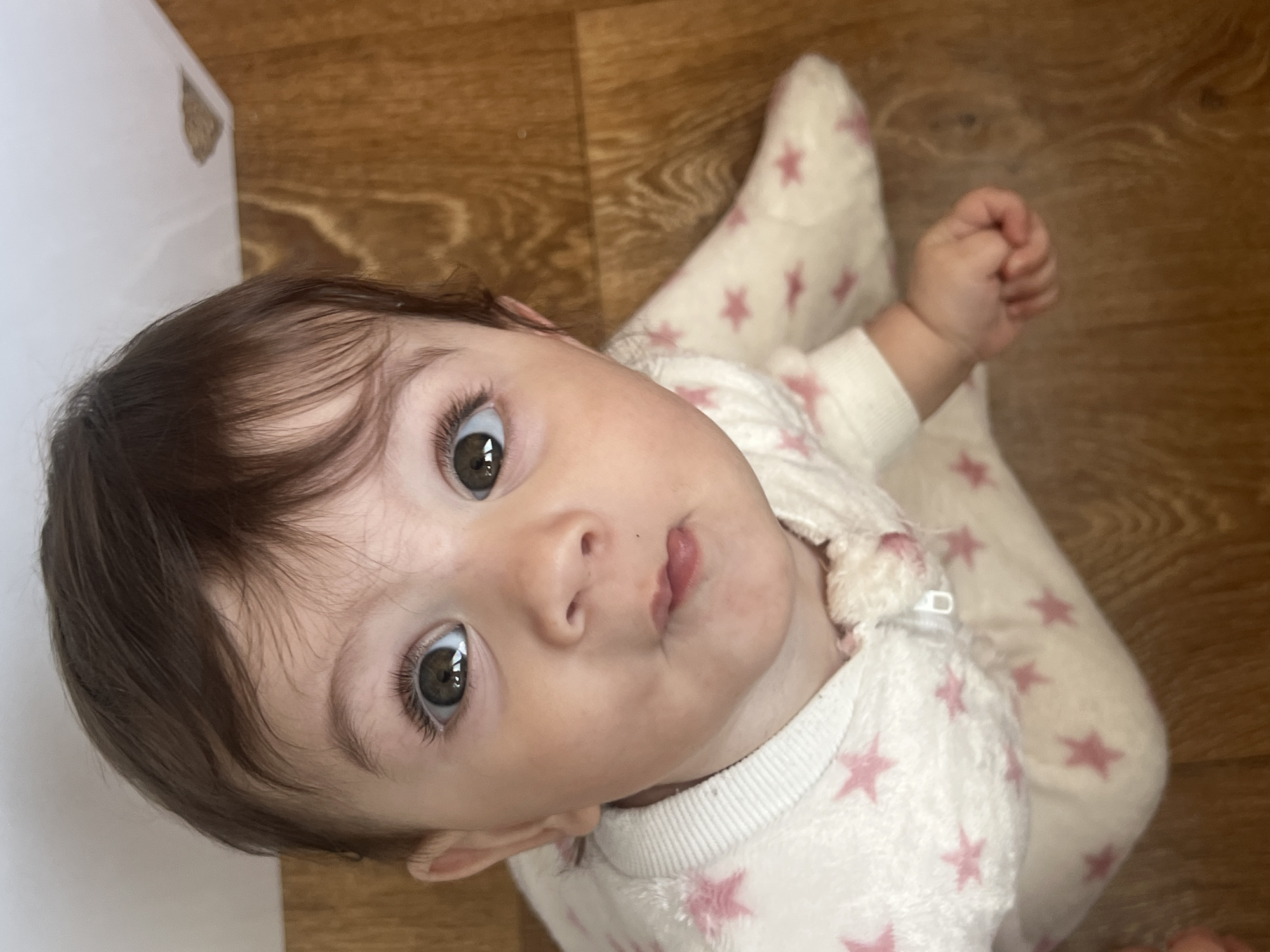 A baby wearing a white onesie with pink stars is lying on a brown, wood-patterned floor. The baby has brown hair and eyes and is looking up at the camera with a slightly puckered expression. The baby's right hand is clenched into a fist. A white wall is visible in the upper left corner of the frame. This image does not provide enough information to determine a specific location in Elena, Bulgaria, or any other city.