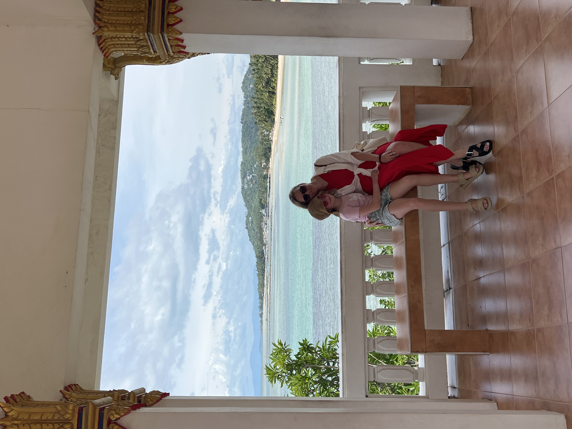 A woman and a young girl are seated on a wooden bench in a covered structure, with a scenic view of the sea and coastline of Baan Hua Thanon, Thailand, in the background. The woman, on the right, is wearing a red dress, a white cover-up, and sunglasses. The girl, on the left, is wearing a pink t-shirt, denim shorts, and a straw hat. They are posed closely together, with the woman's arm around the girl. The bench is made of wood and is positioned on a tiled floor. The structure has ornate pillars with gold detailing on the sides. Beyond the bench is a white railing, and further in the distance, the turquoise sea meets a sandy beach lined with palm trees and houses. The sky is partly cloudy, with a mix of blue and white.