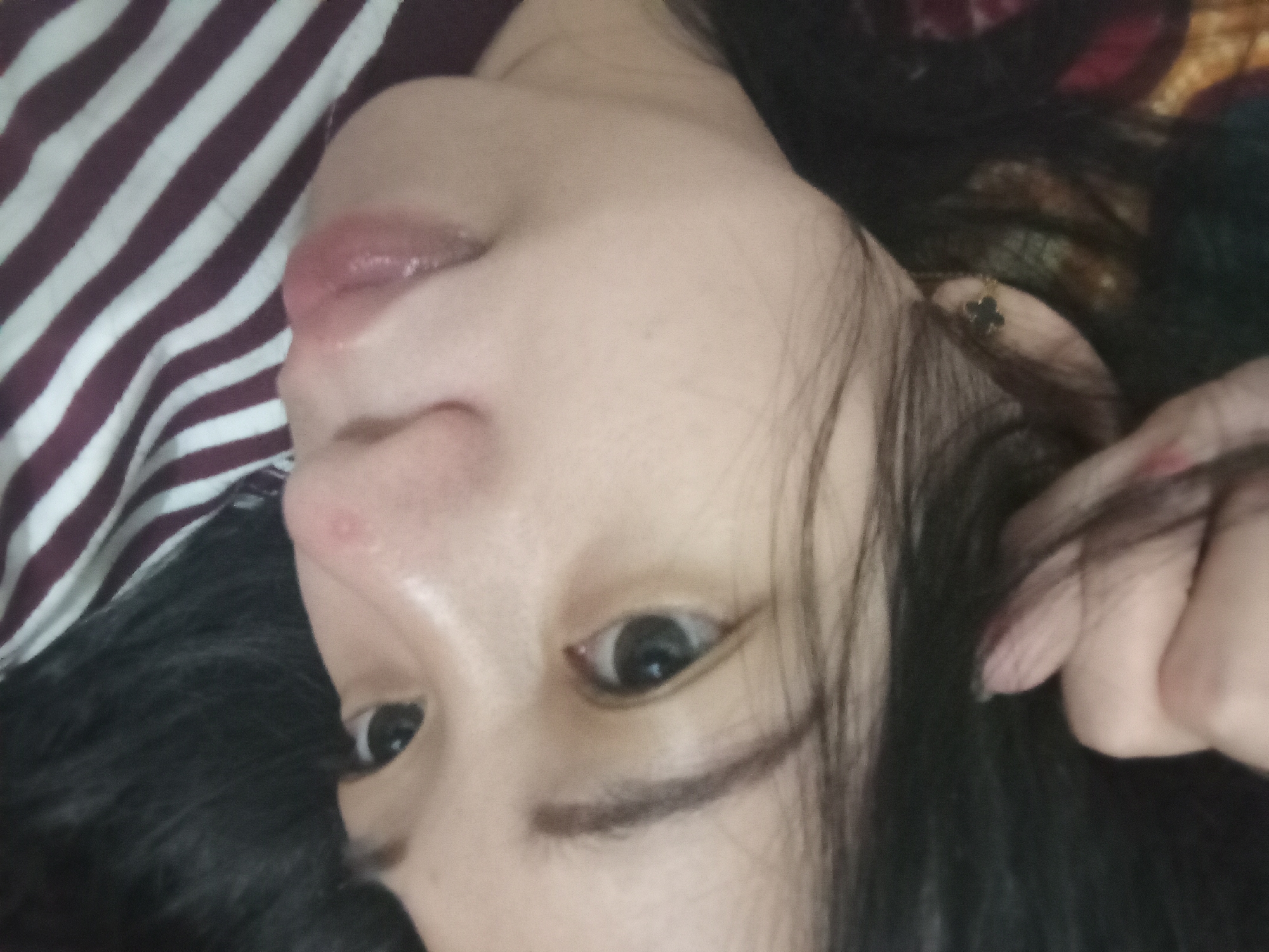 The image is a close-up, self-taken photograph of a young woman's face, angled downwards. She has dark hair, fair skin, and is wearing a striped shirt. A hand is visible near her head, with painted fingernails. The overall lighting is soft. No specific location details are discernible from the image.