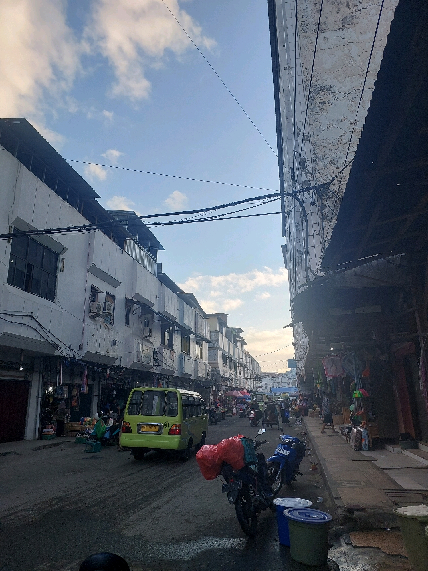 This image captures a street scene in Ambon, Indonesia, likely a marketplace area, with buildings lining both sides of a road. The architecture suggests a mix of commercial and residential use, with multiple stories and visible air conditioning units on the buildings to the left. The street itself is paved and appears wet in some areas, possibly from recent rain or cleaning.
Several vehicles are present: a bright yellow minivan with a license plate visible, several motorcycles, and a larger truck further down the street. The motorcycles are parked along the sidewalk and in the road, some carrying cargo, including red plastic bags. People are visible walking, standing, and interacting within the scene. Some appear to be vendors or shoppers. Goods are displayed along the sidewalks, hanging from stalls or piled on the ground, suggesting active commerce. Overhead, numerous electrical wires crisscross the sky, typical of urban infrastructure. The sky is partly cloudy with hints of a sunset glow, indicating it's likely late afternoon or early evening. The overall impression is a bustling, everyday street scene in a Southeast Asian city.