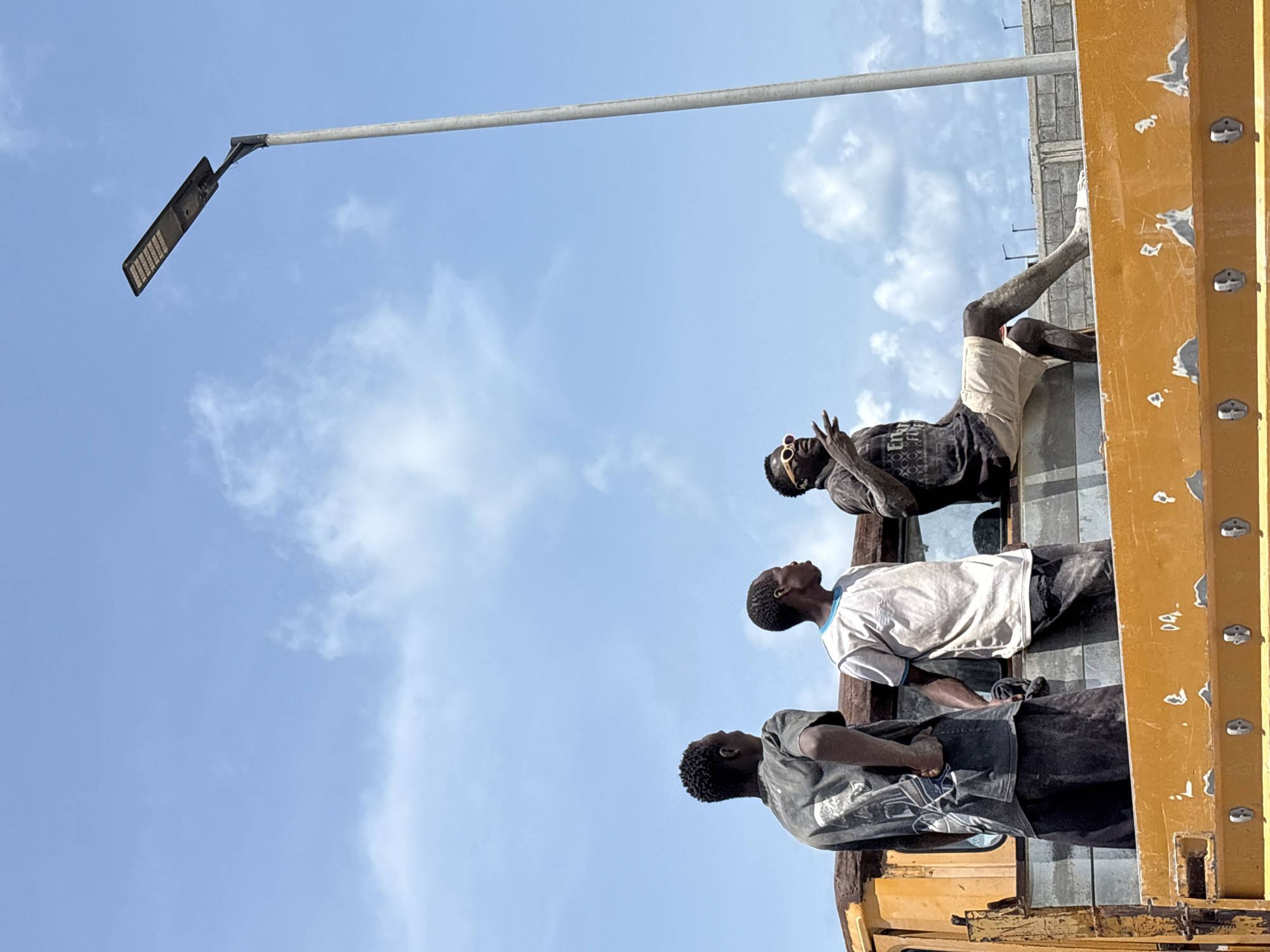 This image captures an outdoor scene in Igboefon, Nigeria, featuring four men on a large yellow structure, likely a truck bed or a similar platform, beneath a bright blue sky with scattered white clouds. The strong sunlight suggests it is daytime with fair weather.
In the foreground, one man lies relaxed on his back on the yellow surface, wearing light-colored shorts, a dark t-shirt, and sunglasses. His skin and clothing are visibly covered in dust, and one arm is casually raised. Behind him, two other men stand looking upwards. They are also covered in dust, one wearing a light t-shirt and the other a darker one. A fourth man, similarly dusty and in a dark shirt, is partially visible to the left, also gazing upwards. Their postures suggest they might be resting or observing something above them.
A prominent grey street light pole extends horizontally across the upper portion of the frame, with a modern, rectangular LED light fixture attached to its end. In the background, behind the men and the yellow structure, part of a building constructed with concrete blocks is visible. The yellow structure itself shows signs of wear and has various metal fixtures or bolts on its side. There is no discernible text visible in the image. The scene conveys a moment of pause or observation in a dusty, working environment.