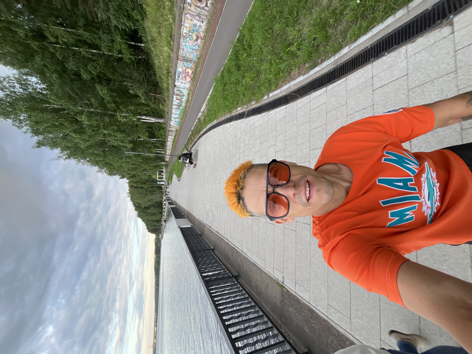 A man with bright orange hair and sunglasses is taking a selfie outdoors along a waterfront promenade. He is wearing an orange t-shirt with a teal and white logo that reads "MIA" in large letters, and what appears to be dark pants. He is smiling at the camera.
The setting is a paved walkway beside a body of water, possibly a river or lake, with a metal railing separating the walkway from the water. Beyond the railing, there are trees and a grassy embankment. In the background, there is a path with graffiti art on a wall and some people walking. The sky is overcast, suggesting it might be late afternoon or early evening. The overall scene conveys a casual and relaxed atmosphere. The location is identified as Chaikovsky, Russia.